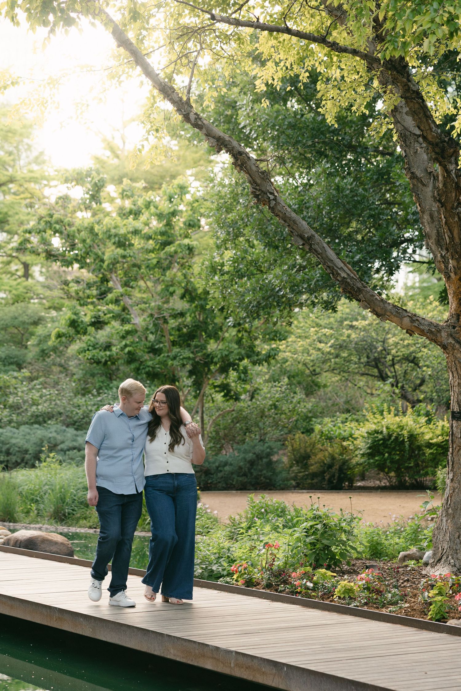Couple walking hand-in-hand on a wooden boardwalk surrounded by lush green gardens in warm sunlight.