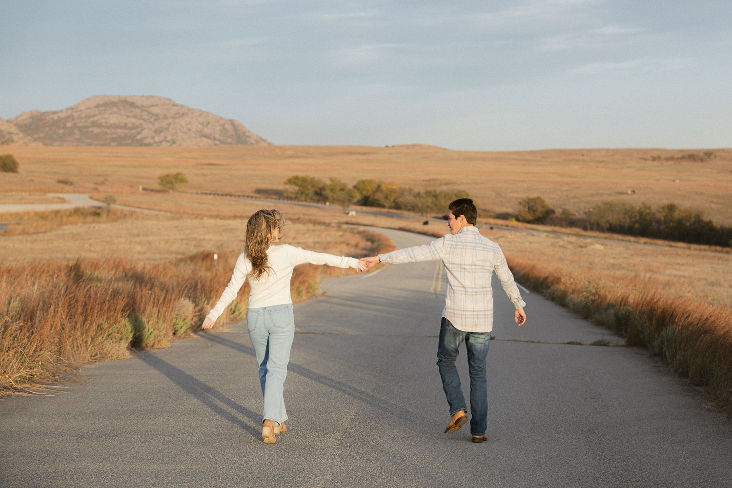 Two people hold hands and walk down a rural road at sunset while wearing casual clothing and facing away from camera.