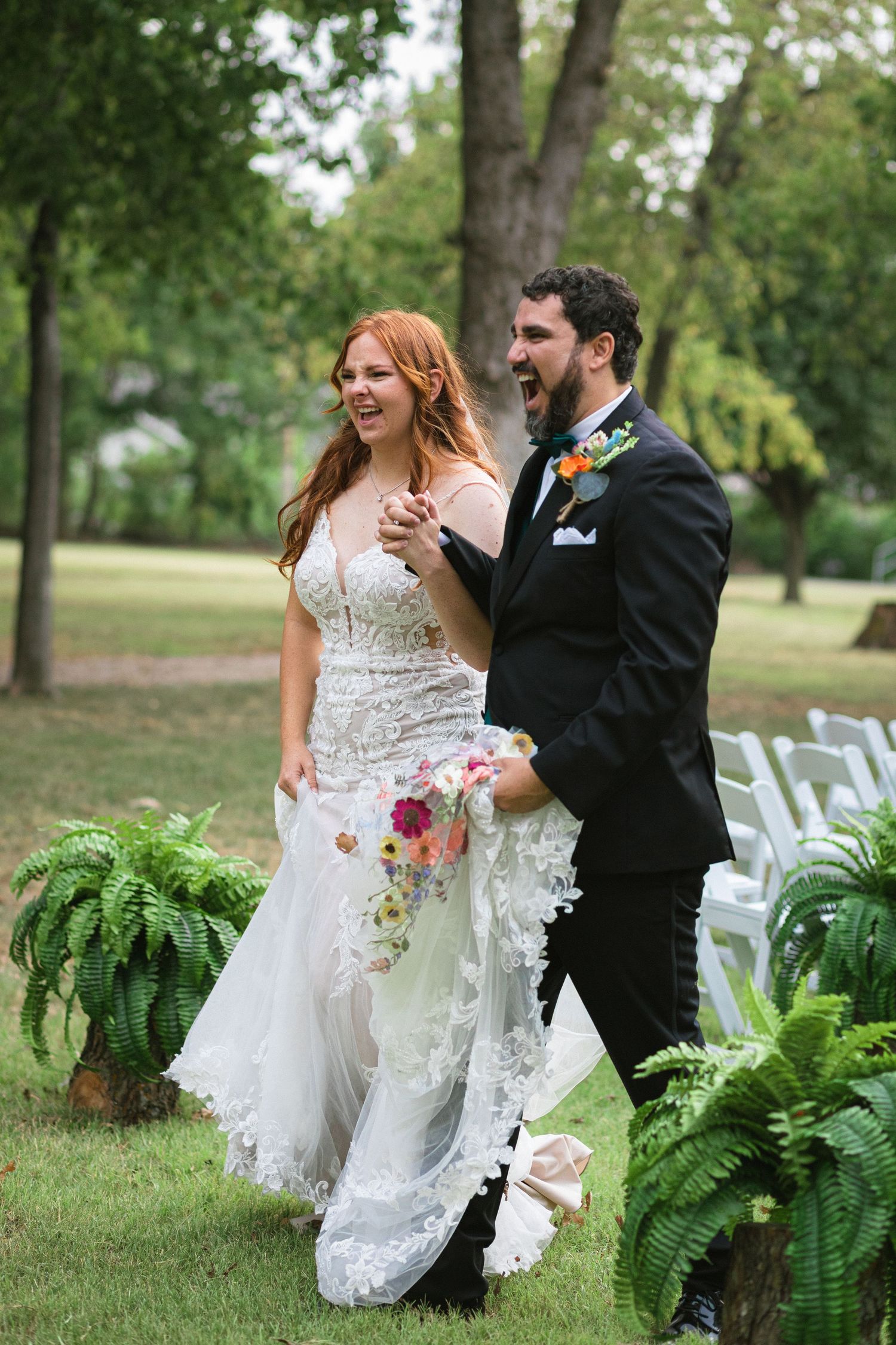 A couple in wedding attire shares joyful moments among lush green ferns during their outdoor garden ceremony.