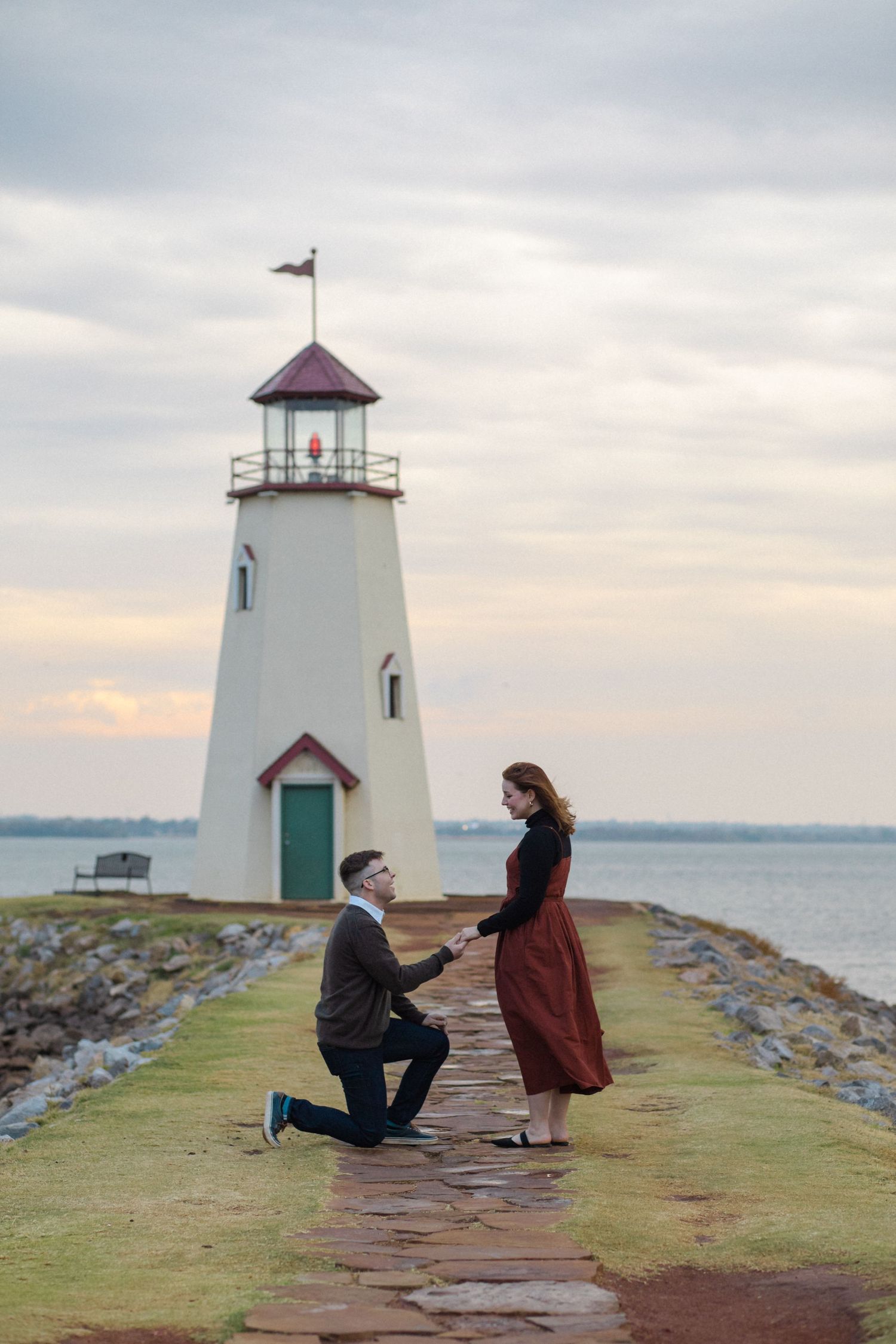 A romantic marriage proposal takes place on a stone walkway leading to a white lighthouse at sunset.