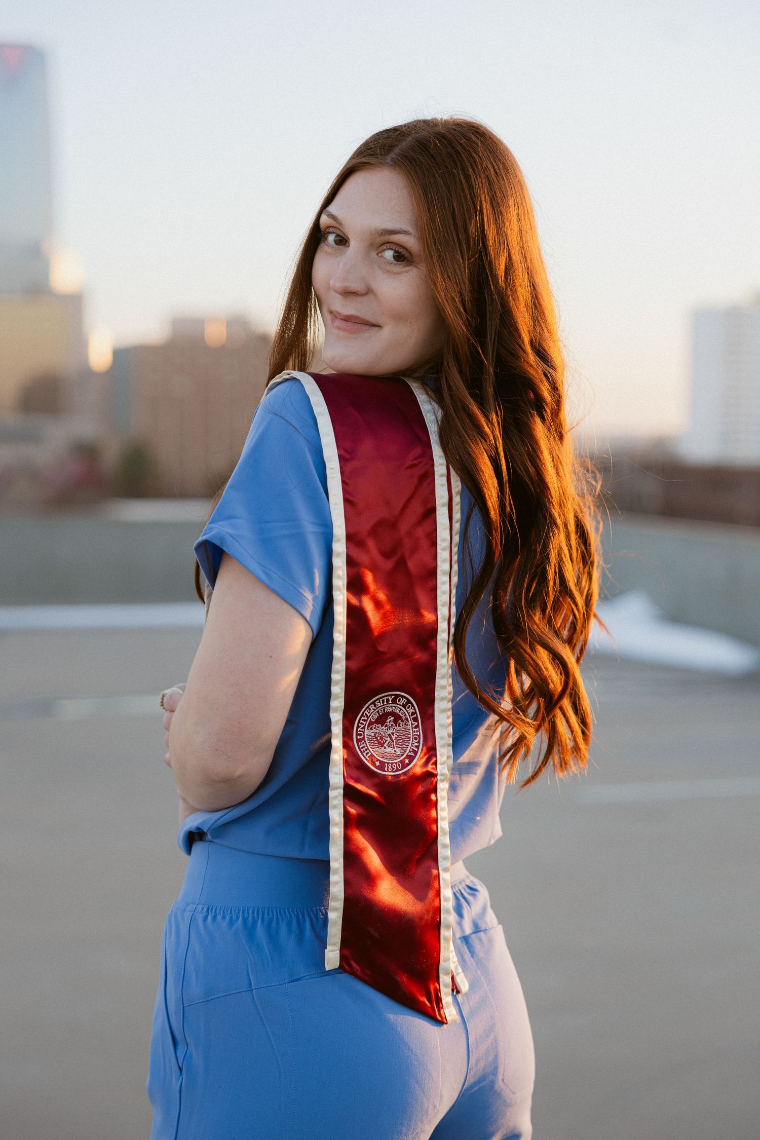 A portrait of someone in graduation attire with blue robes and a red stole against an urban sunset backdrop.
