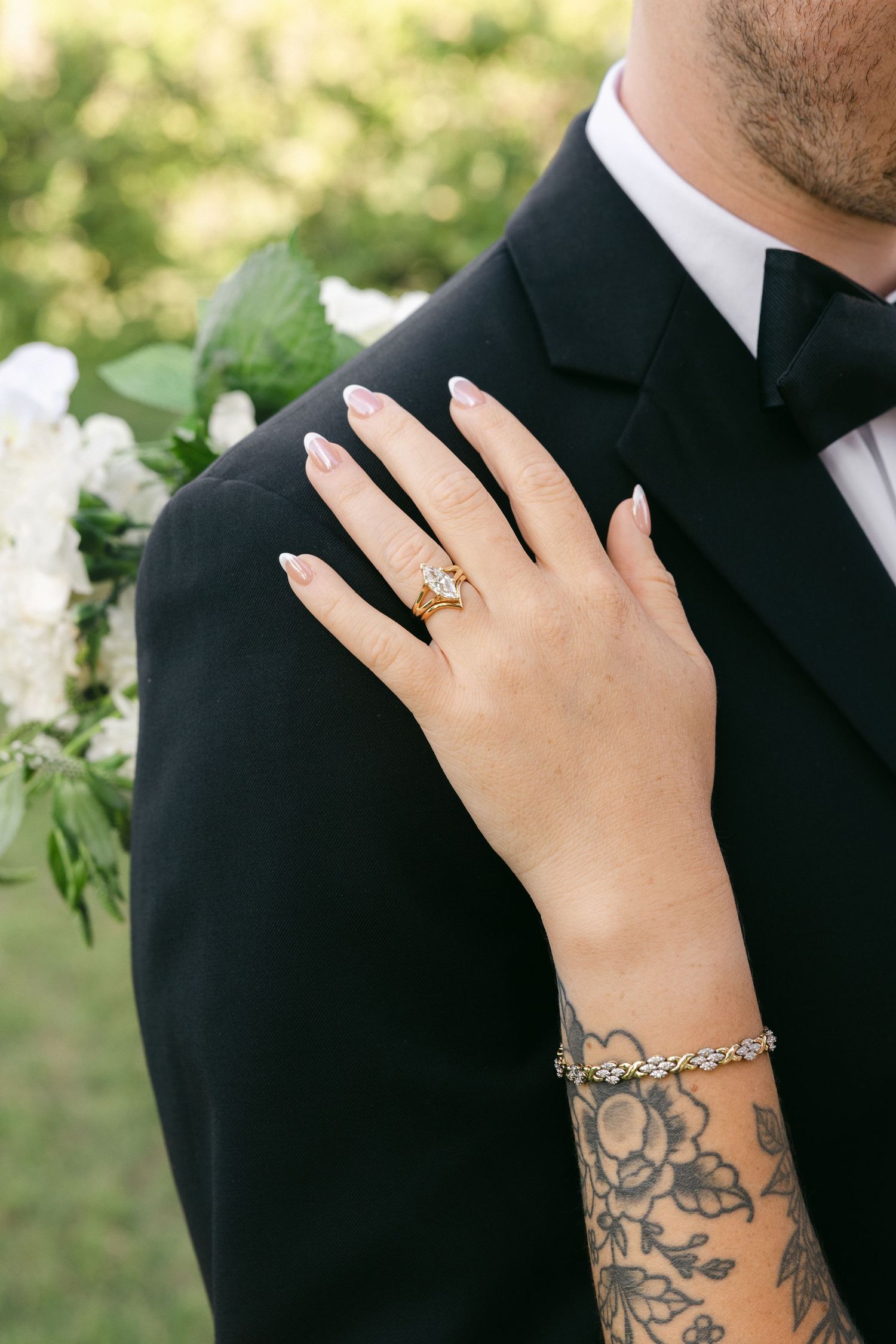 Bride's hand with gold engagement ring and floral tattoo rests on groom's black tuxedo shoulder at wedding.