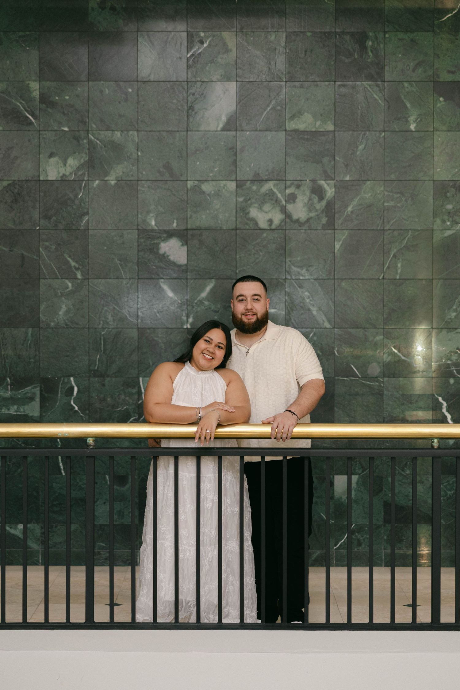 Couple posing elegantly against green marble wall with gold railing, woman in white dress, man in cream shirt.