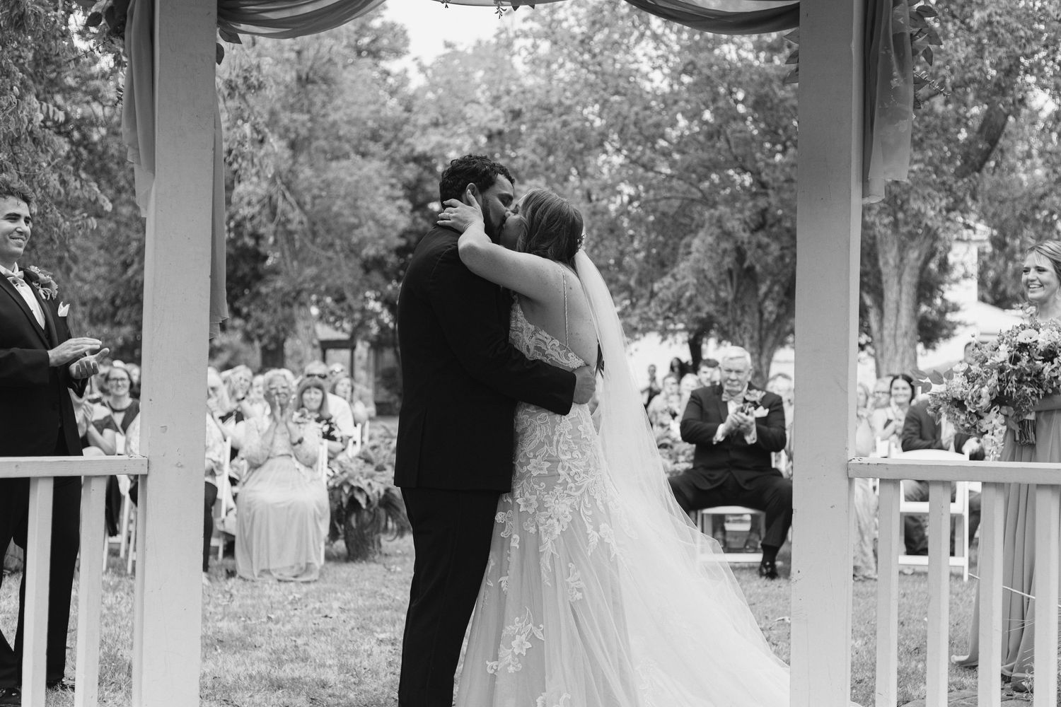 A romantic wedding couple shares their first dance under a white gazebo surrounded by trees and natural light.