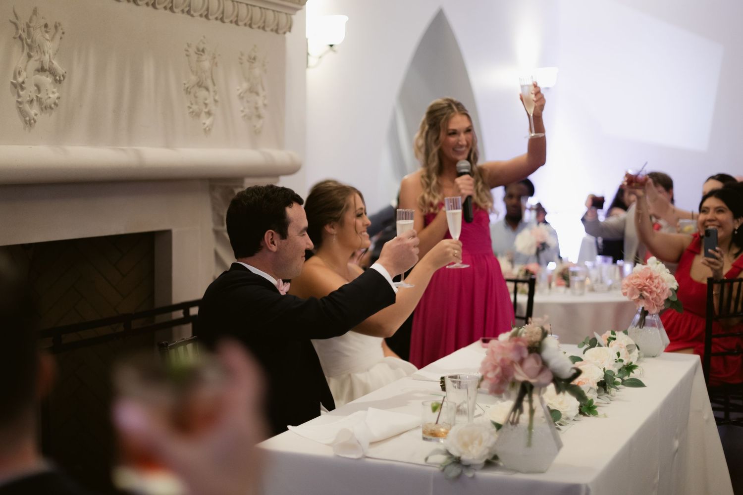 Wedding reception guests raise their glasses in celebration at an elegant indoor venue with white decor.