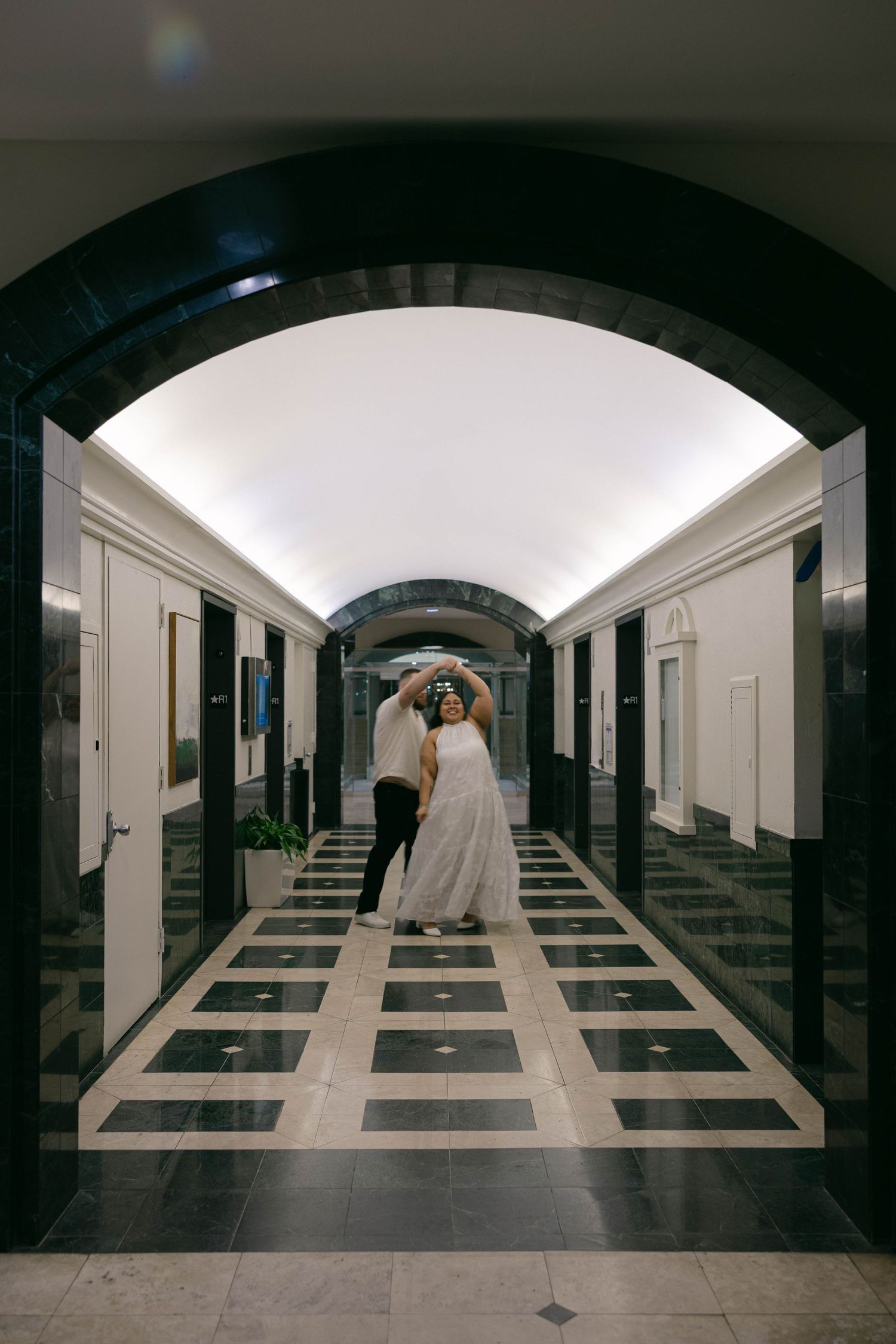 Bride and groom dancing in an elegant hallway with arched ceiling and black-and-white checkered marble floors.