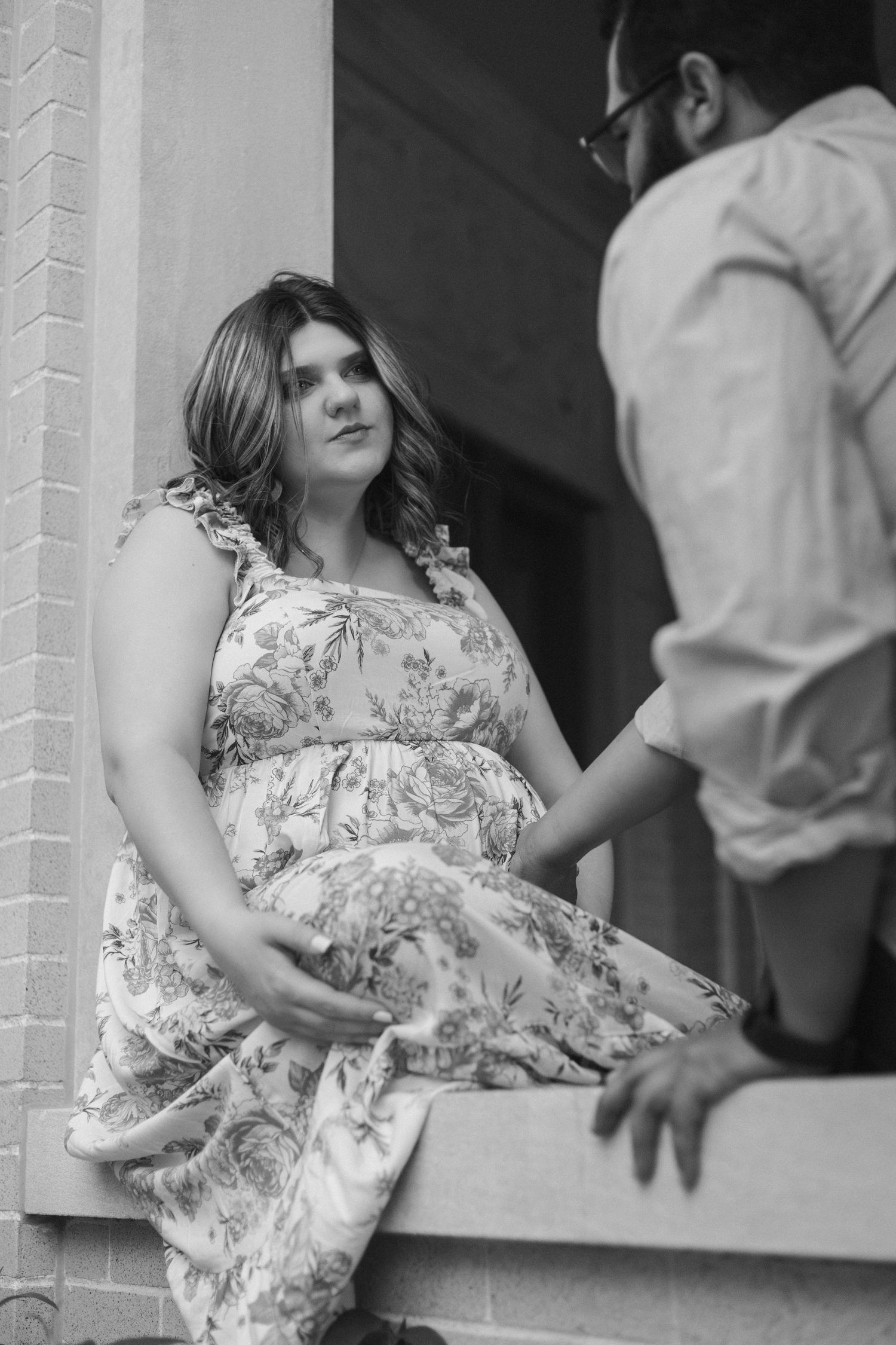 Black and white portrait of an expectant mother in a floral dress sitting on a ledge while interacting with someone beside her.