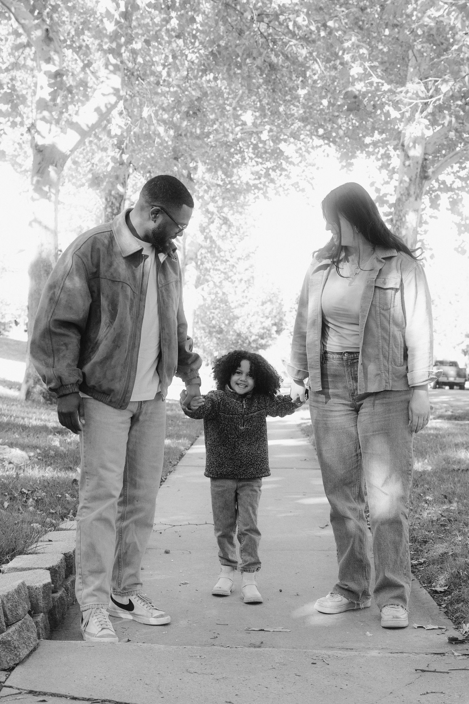 A black and white photograph of a family walking together on a tree-lined path on a sunny day.
