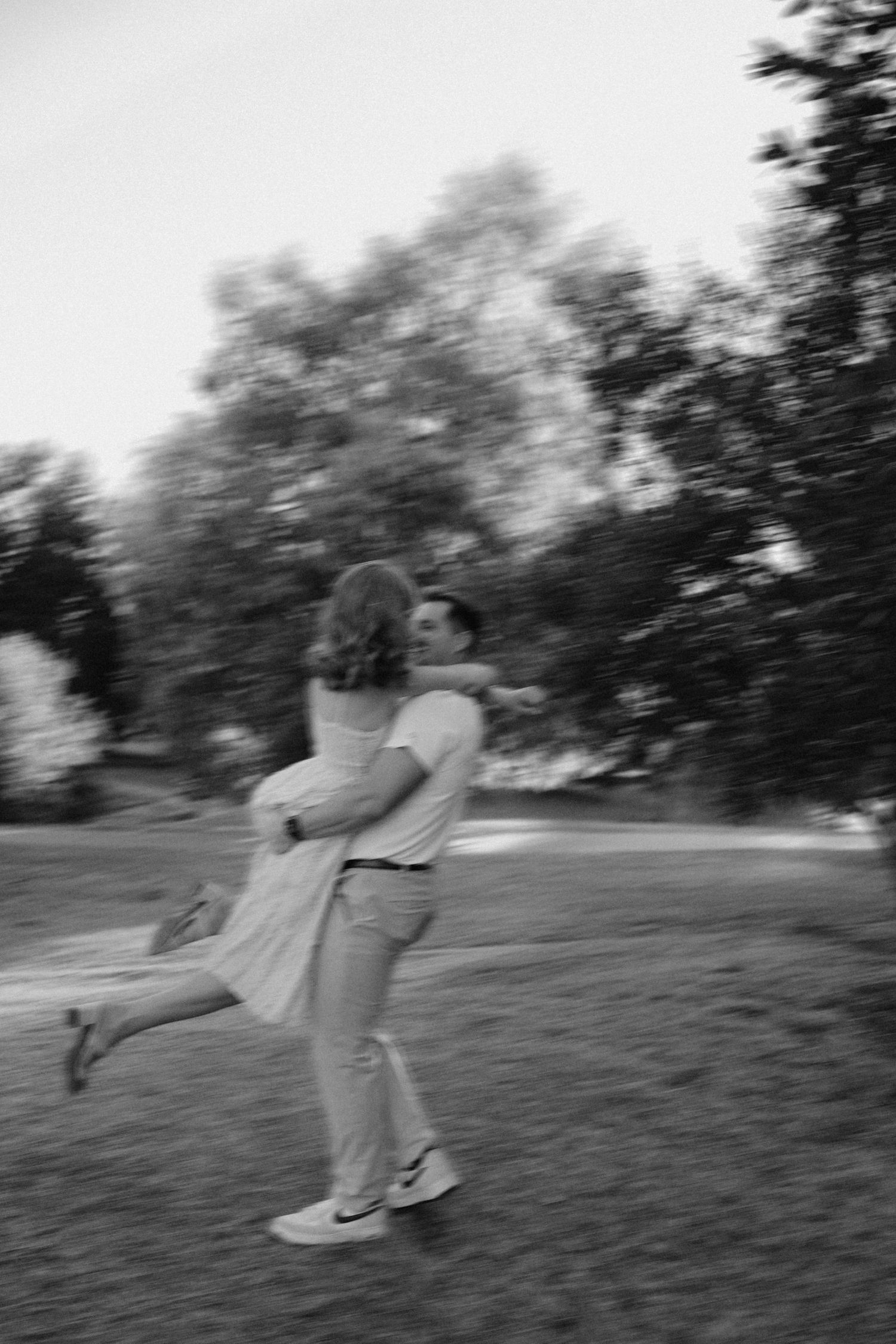 Black and white photo of a couple embracing and spinning joyfully in a park surrounded by trees.