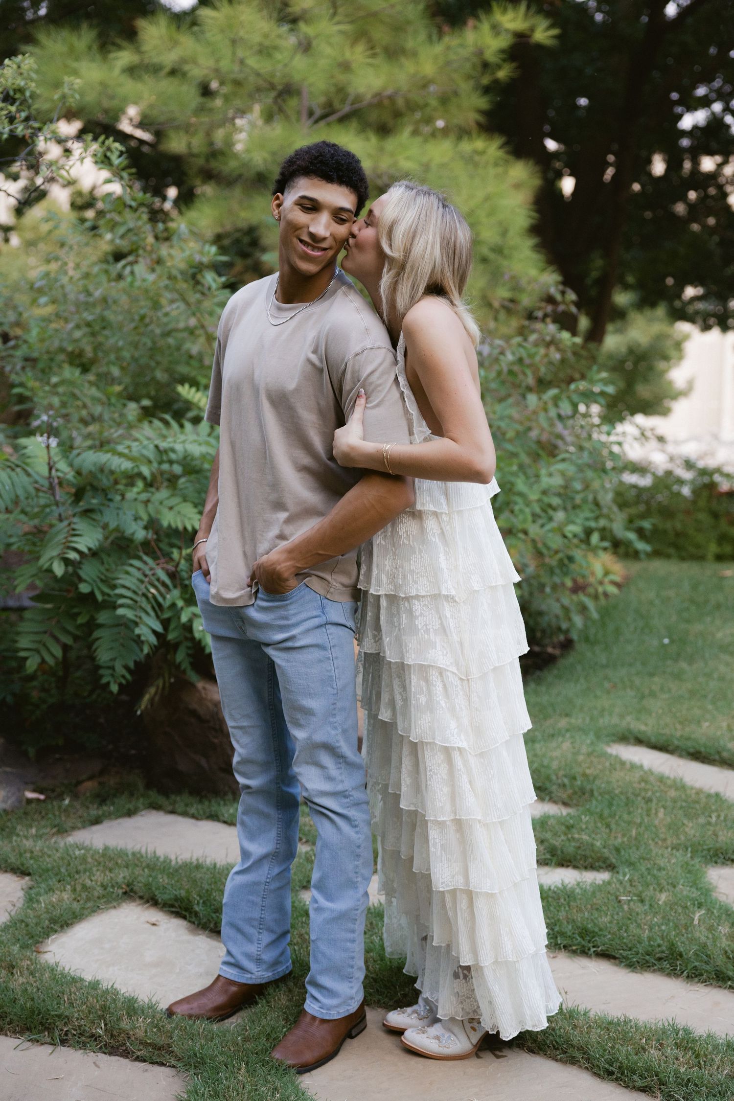 A couple embraces on a garden path, with the woman wearing a tiered white dress and the man in casual attire.