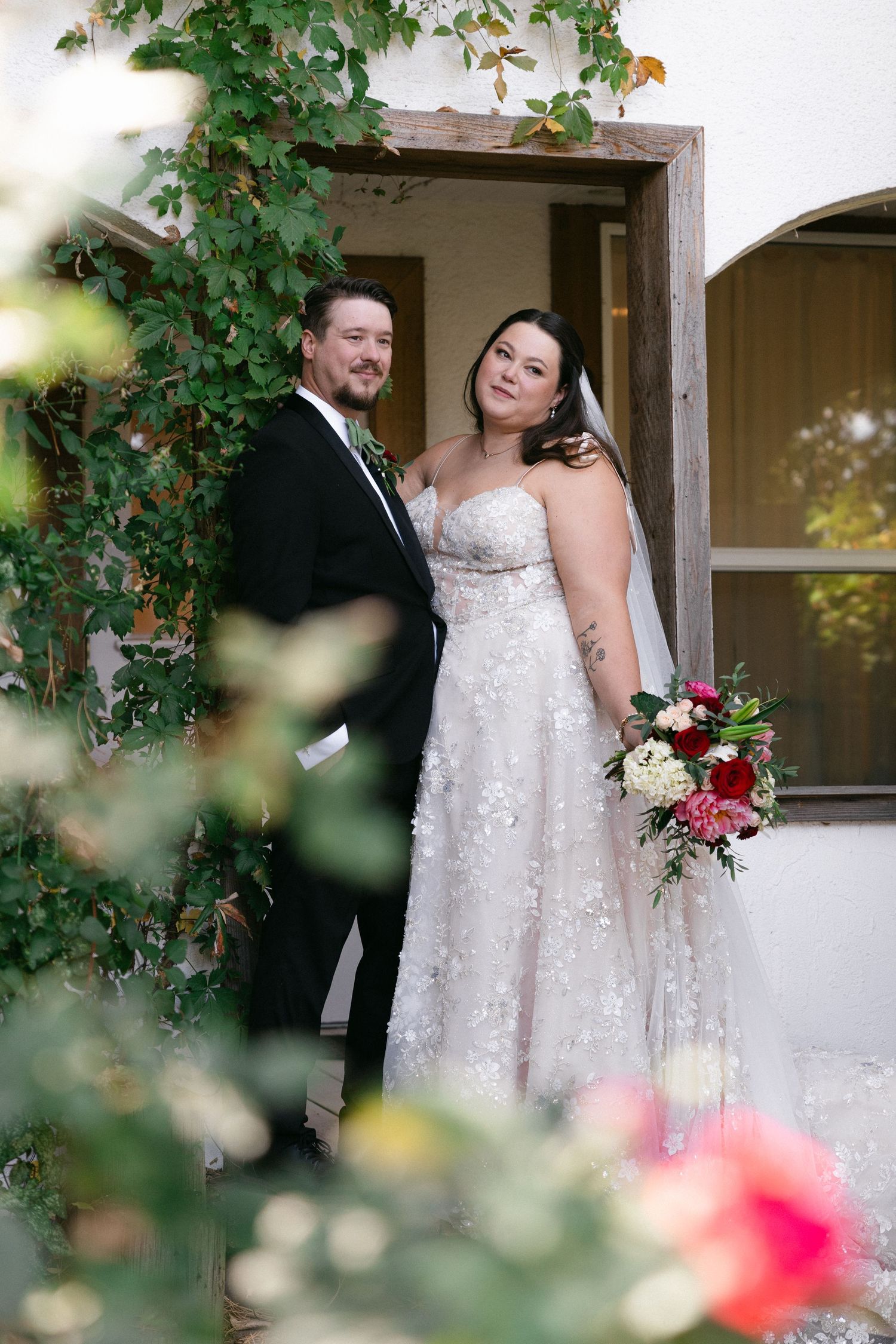 A couple in wedding attire poses together outdoors framed by green ivy and pink flowers.