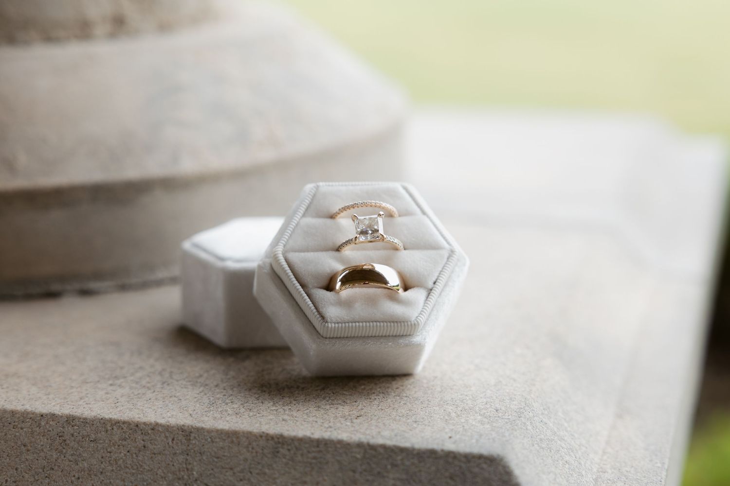 A modern hexagonal white ring box with a gold ring displayed inside sits on a textured gray surface.