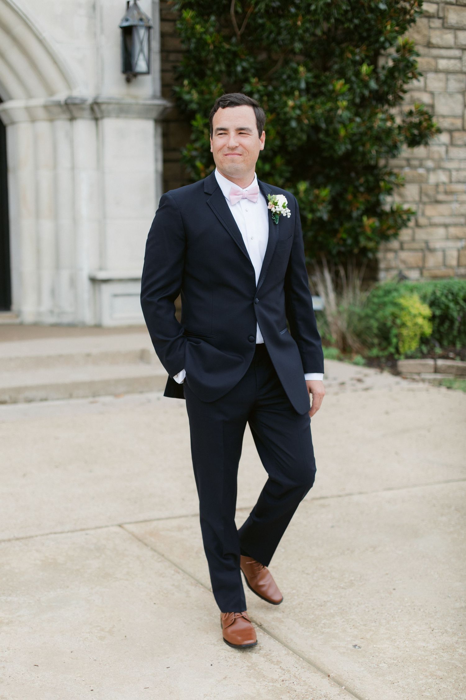 Groom wearing black suit and brown shoes poses playfully in a series of photos against stone building exterior.