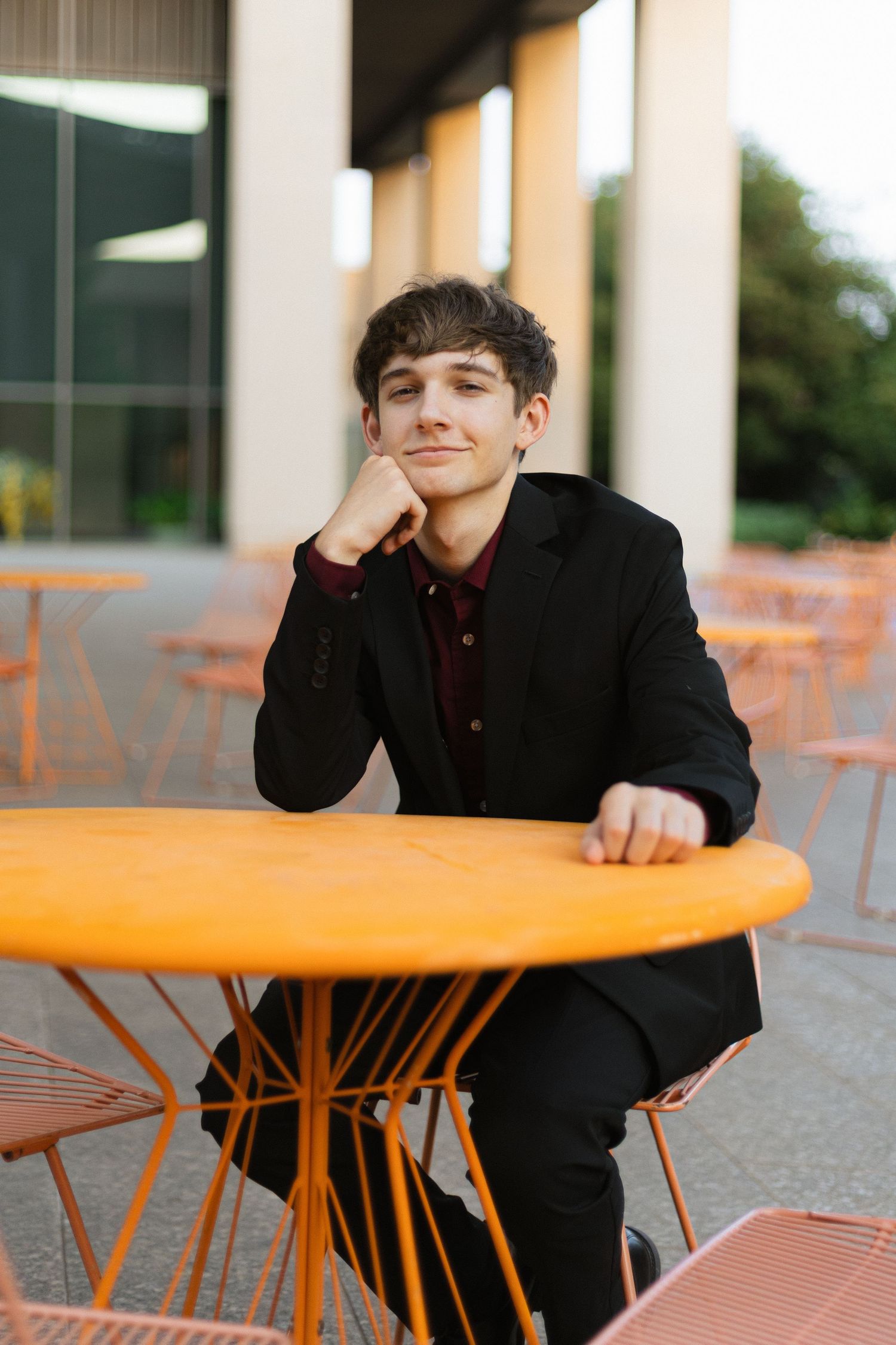 Person in black suit rests at orange outdoor table with contemplative expression.