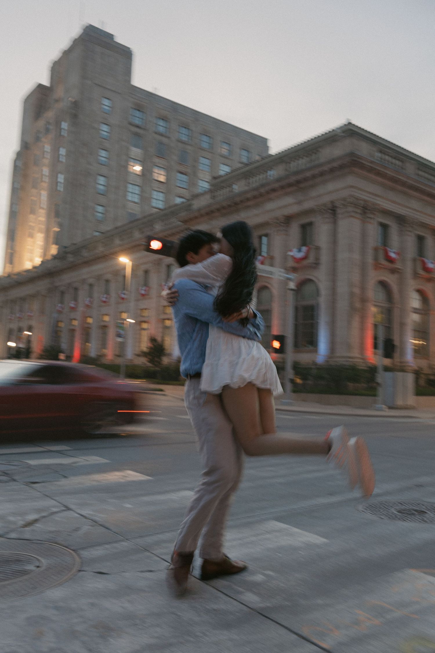 A couple embraces on a city street at dusk, the man lifting the woman as cars and historic buildings blur behind them.