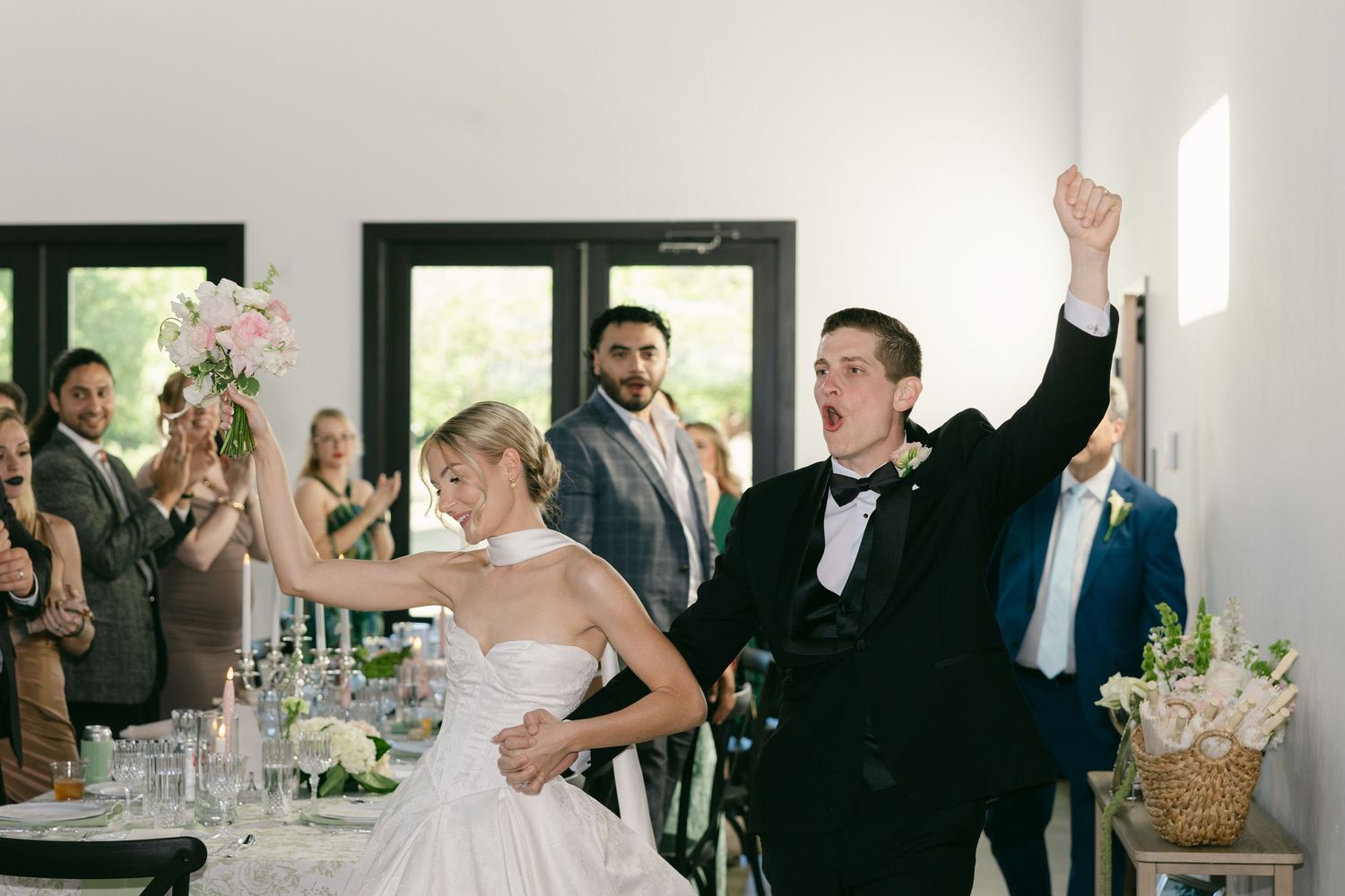 Joyful bride and groom celebrate their wedding reception entrance, raising arms in excitement as guests applaud around decorated tables.