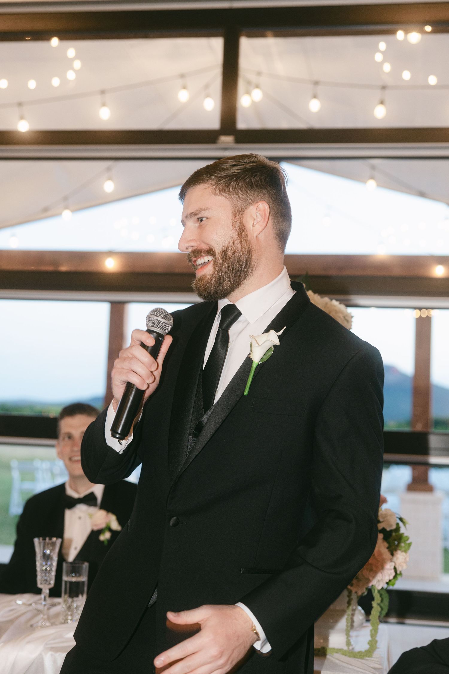 Bearded groomsman in black tuxedo delivering a speech with microphone at wedding reception.