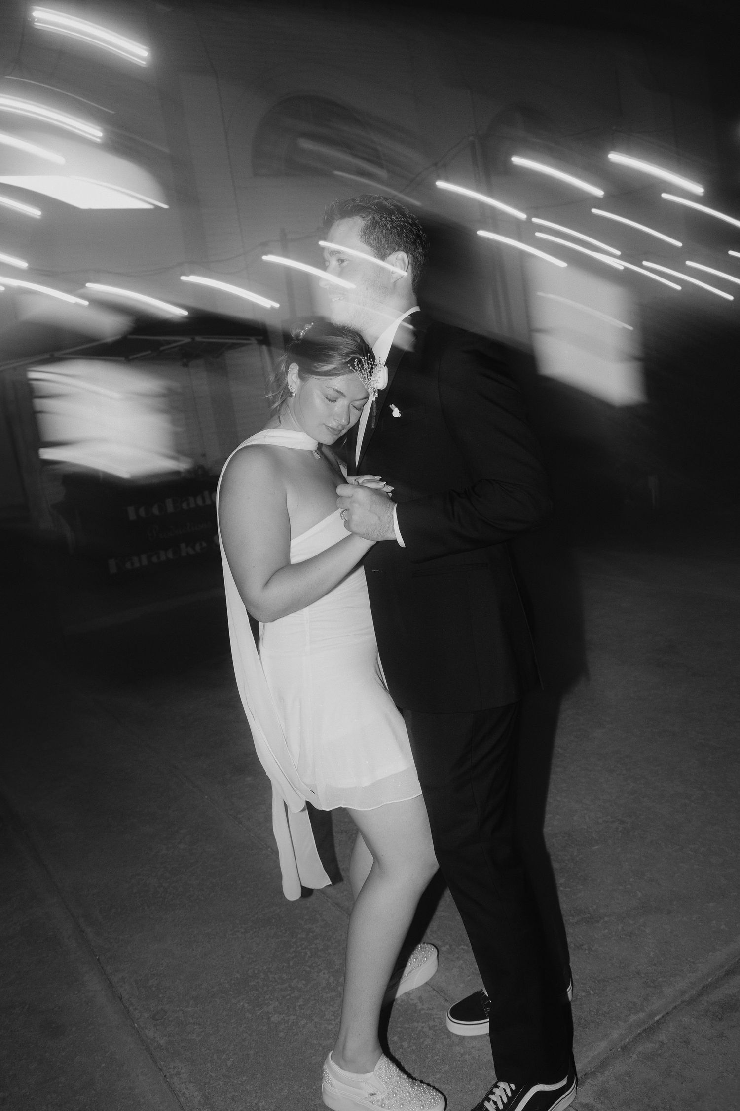 Black and white photo of a couple slow dancing outdoors at night, with motion blur light streaks in the background.
