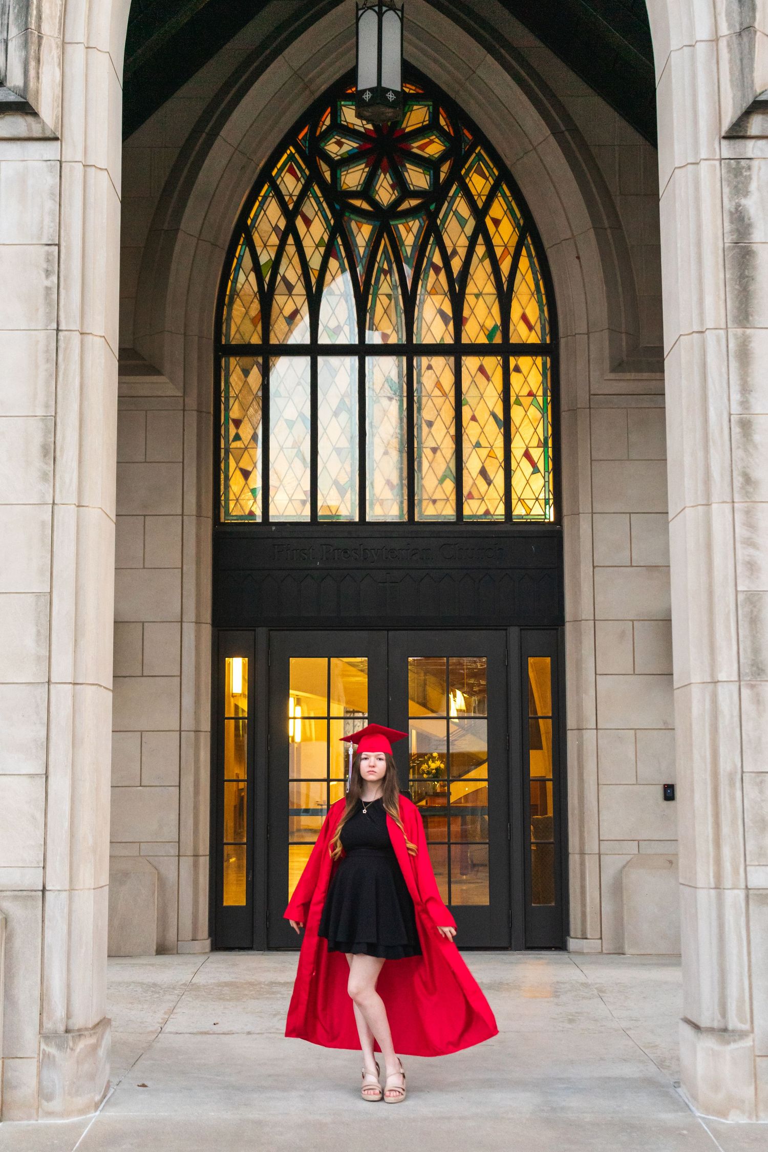 Graduate in red academic regalia poses in gothic archway with stained glass windows.