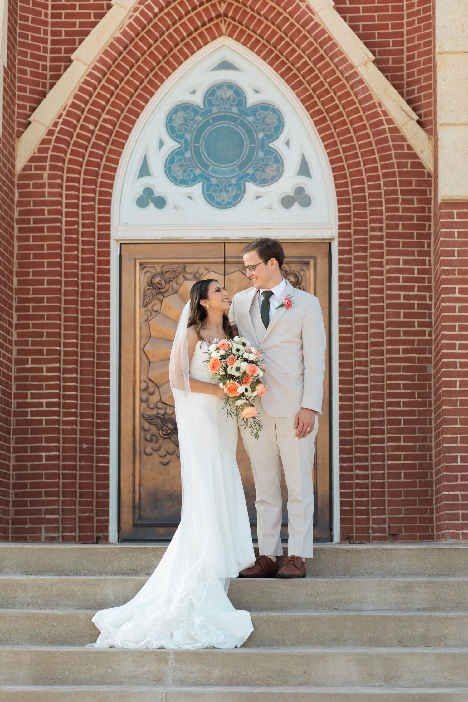Wedding couple poses in formal attire on church steps with ornate archway above.