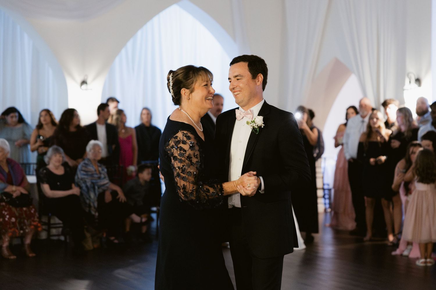Wedding reception guests dancing and socializing in an elegant white-draped venue with ambient lighting.