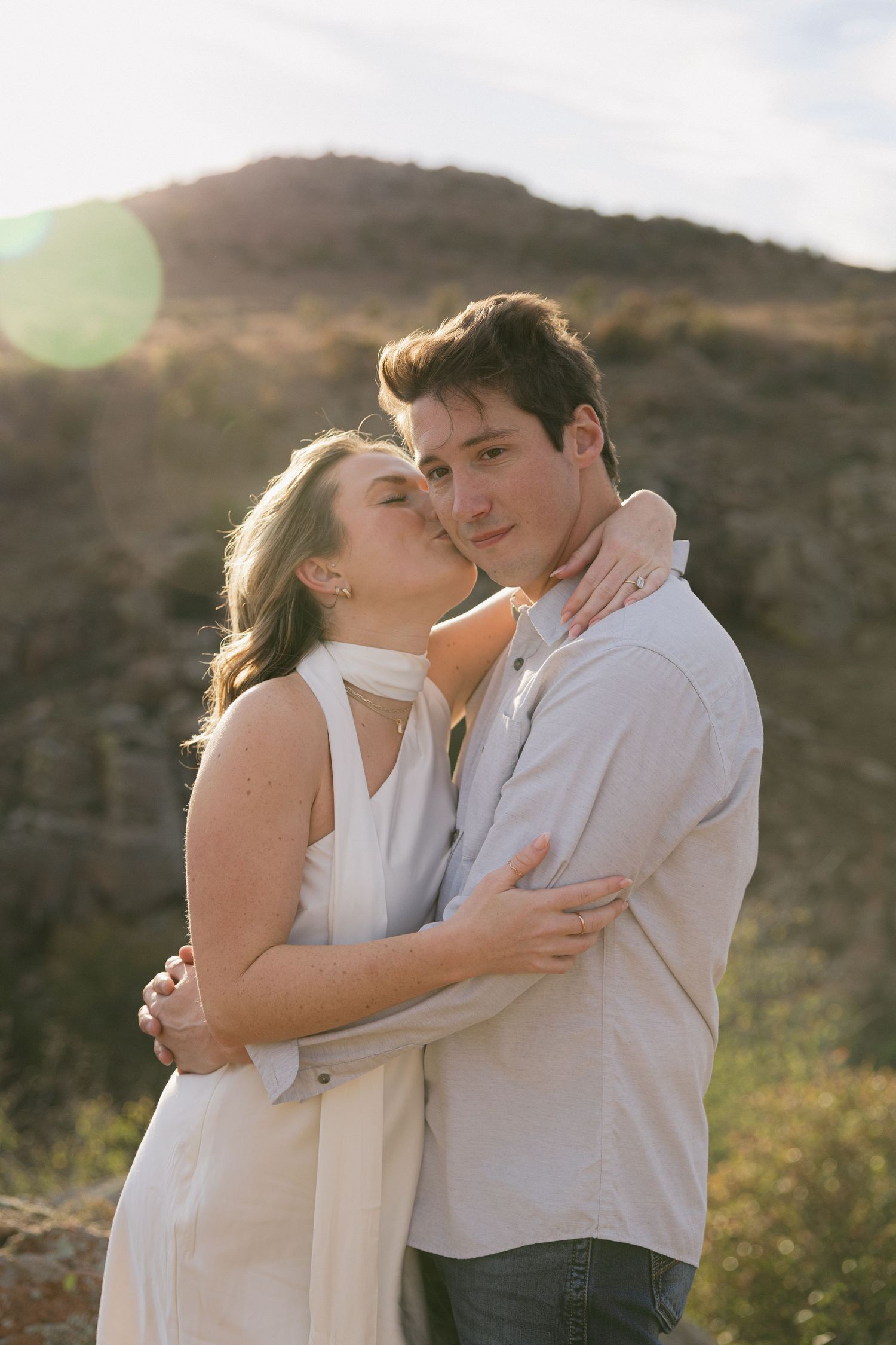 A romantic couple embraces in golden sunlight against a mountainous desert backdrop during sunset.