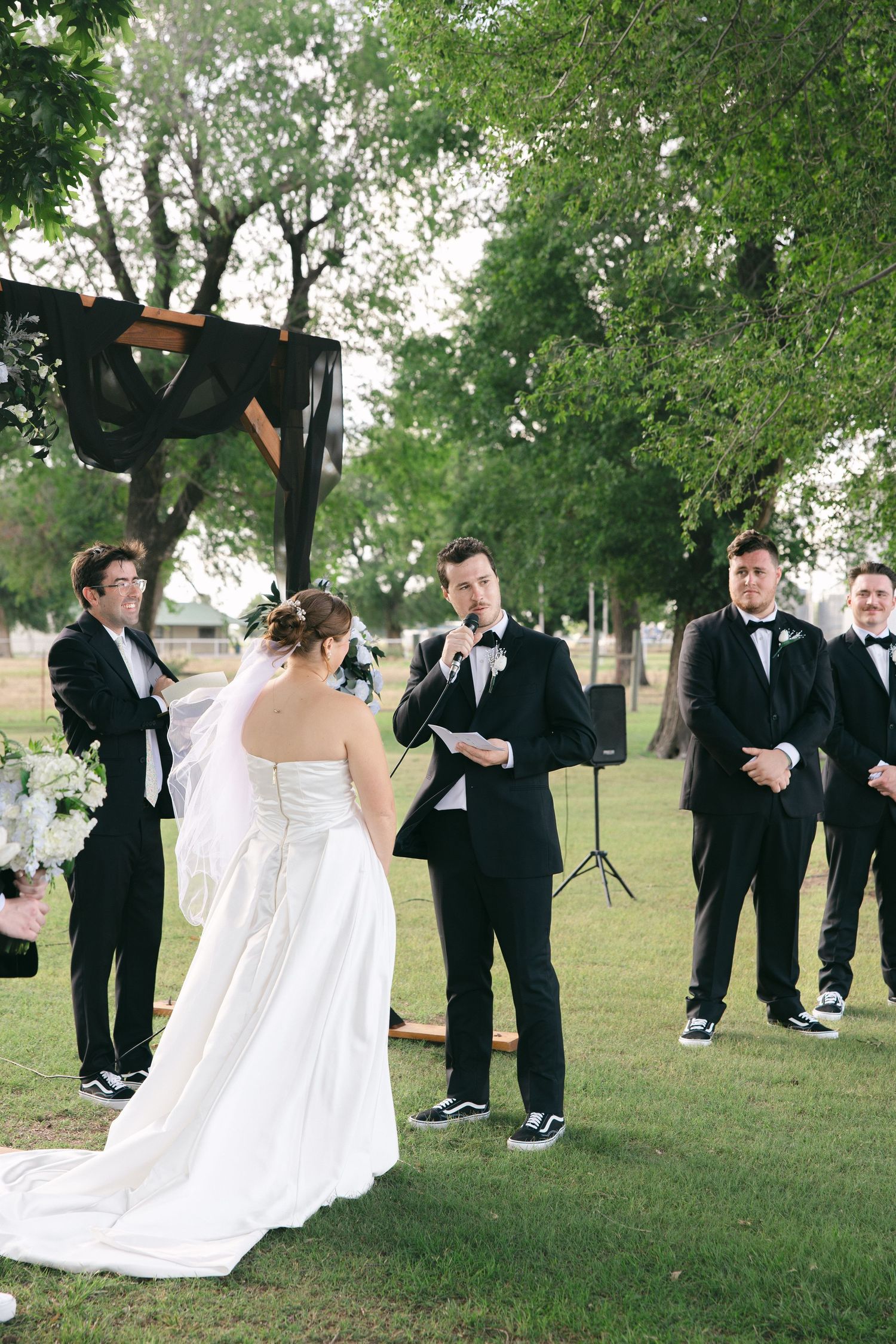 Outdoor wedding ceremony with bride and groom exchanging vows, groomsmen in black tuxedos standing on green lawn.