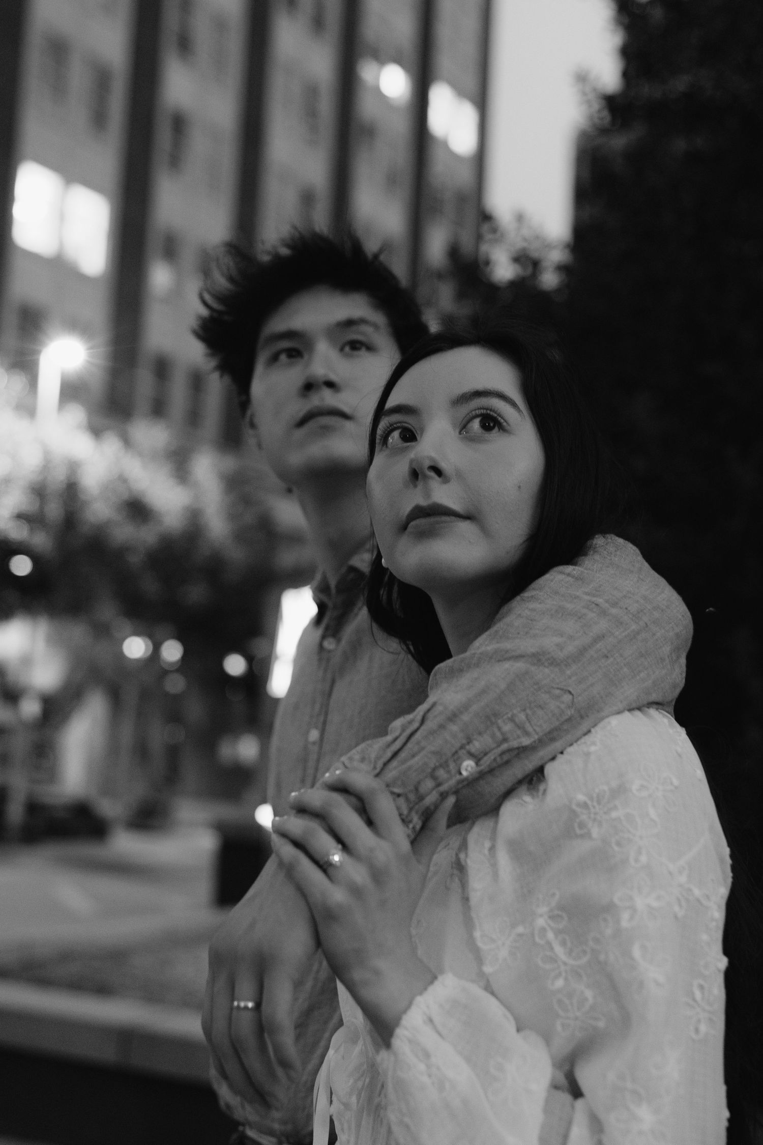 Black and white photo of a couple standing together on a city street at night, woman in white blouse.