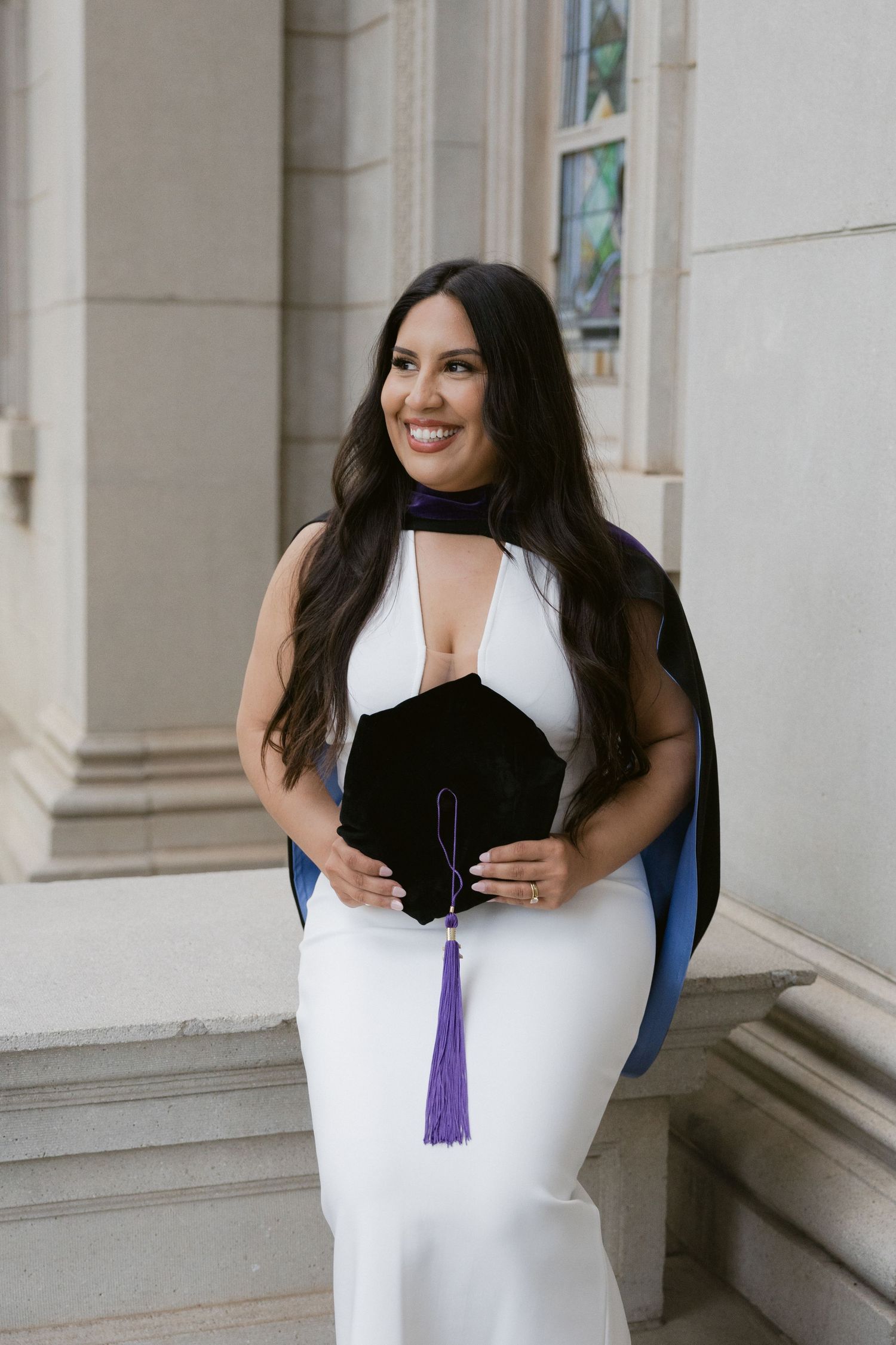 A graduate in a white dress poses for photos with a purple tassel while smiling in a columned hallway.