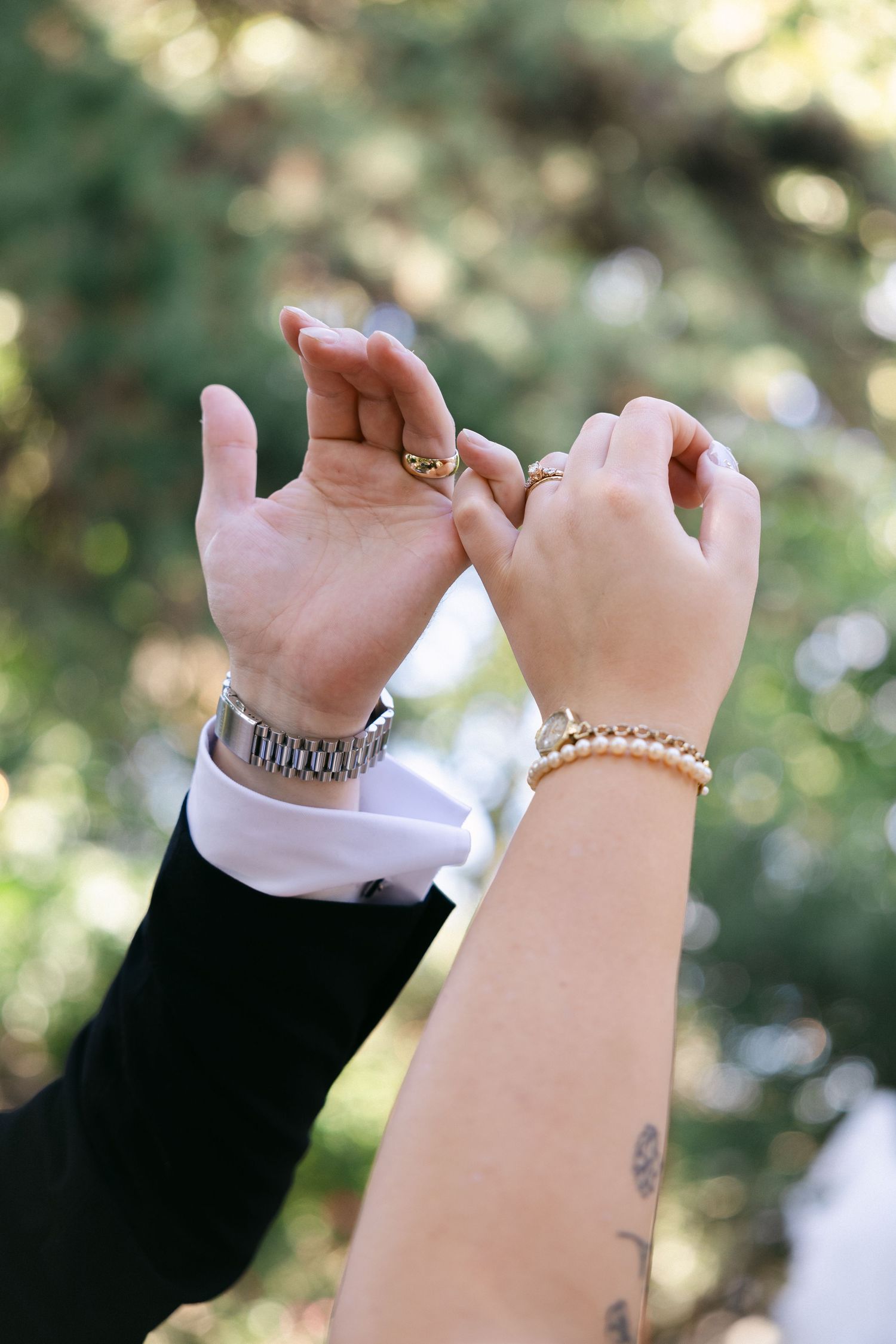Two hands making pinky promise gestures with rings and bracelets against a blurred green outdoor background.