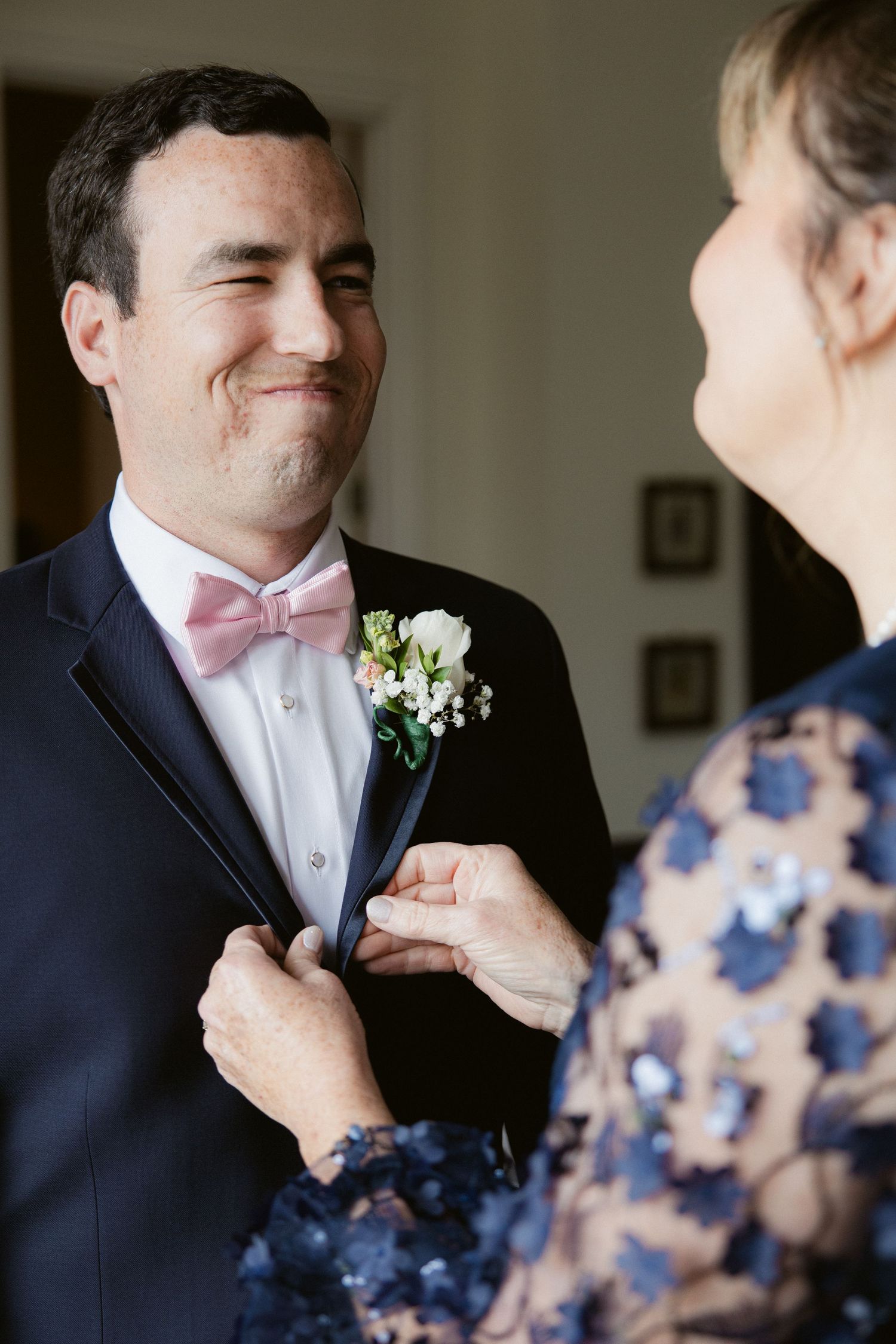 A groom in a navy suit with pink bowtie smiles while someone adjusts his boutonniere before the wedding ceremony.
