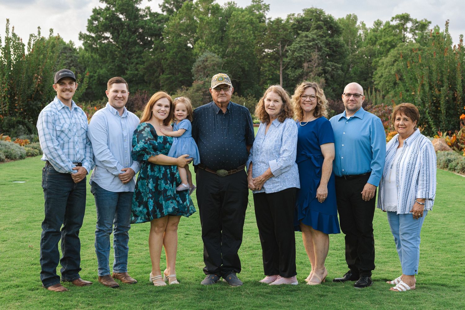 A group portrait of nine people standing together on a manicured lawn with evergreen trees.