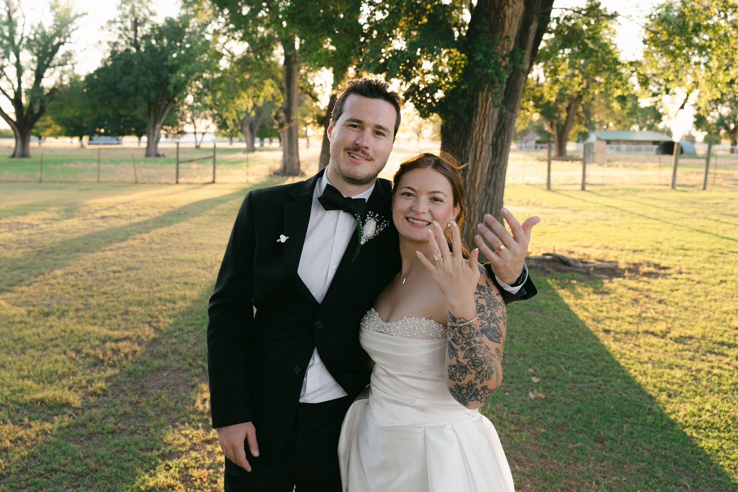Newlywed couple smiling outdoors in wedding attire, bride showing off ring with tattoos, groom in tuxedo, golden hour.