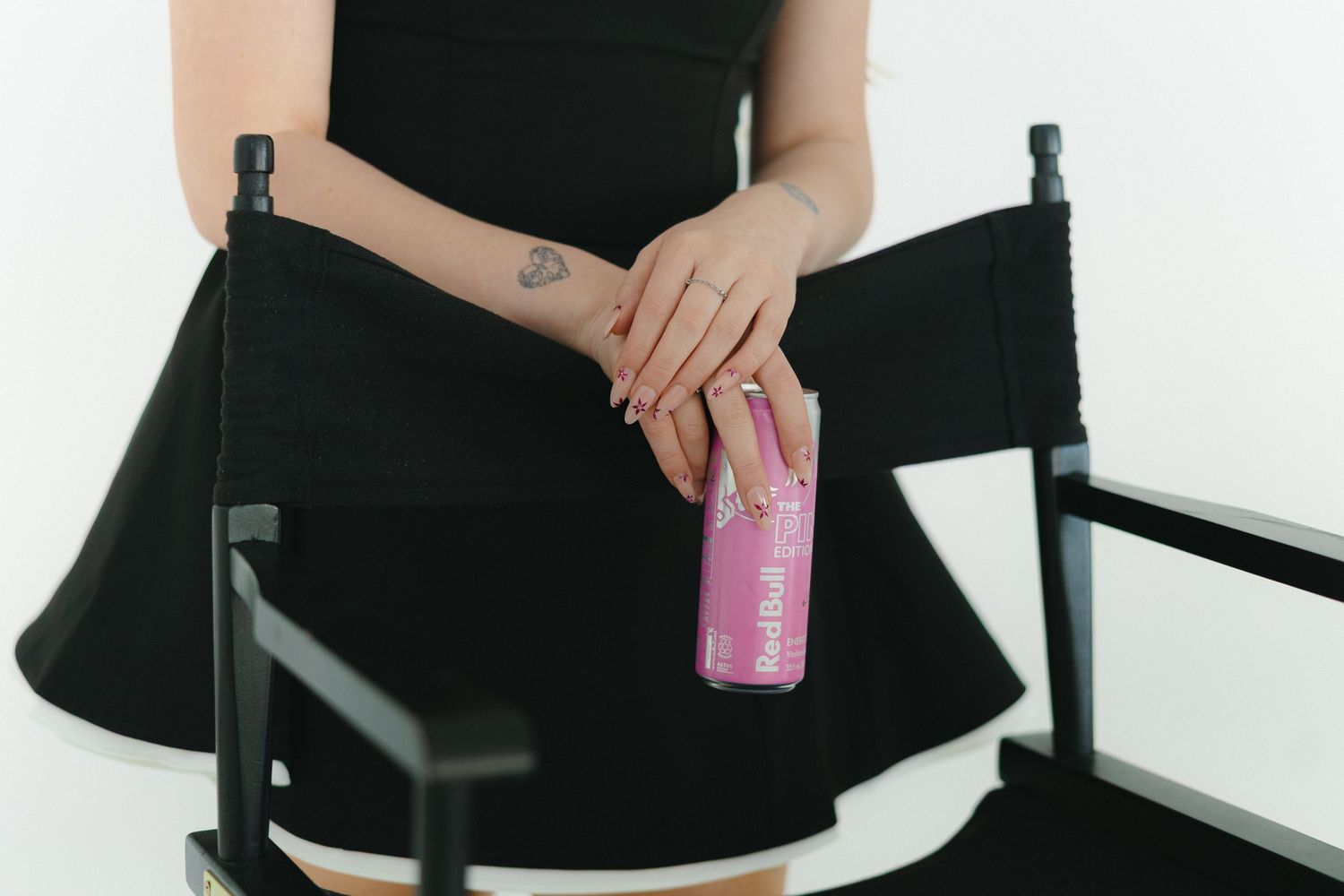 Woman in black dress holding a pink beverage can, showcasing a small tattoo on her wrist.
