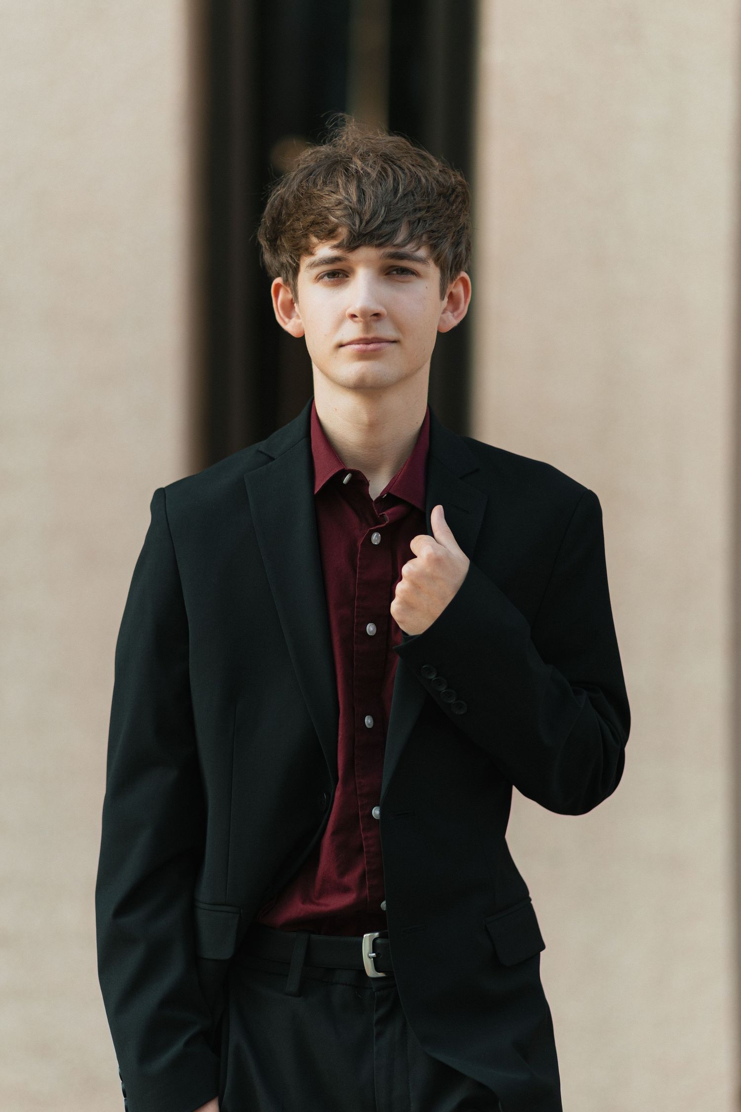 Portrait of a young person in a black blazer and burgundy shirt posing against a light wall.