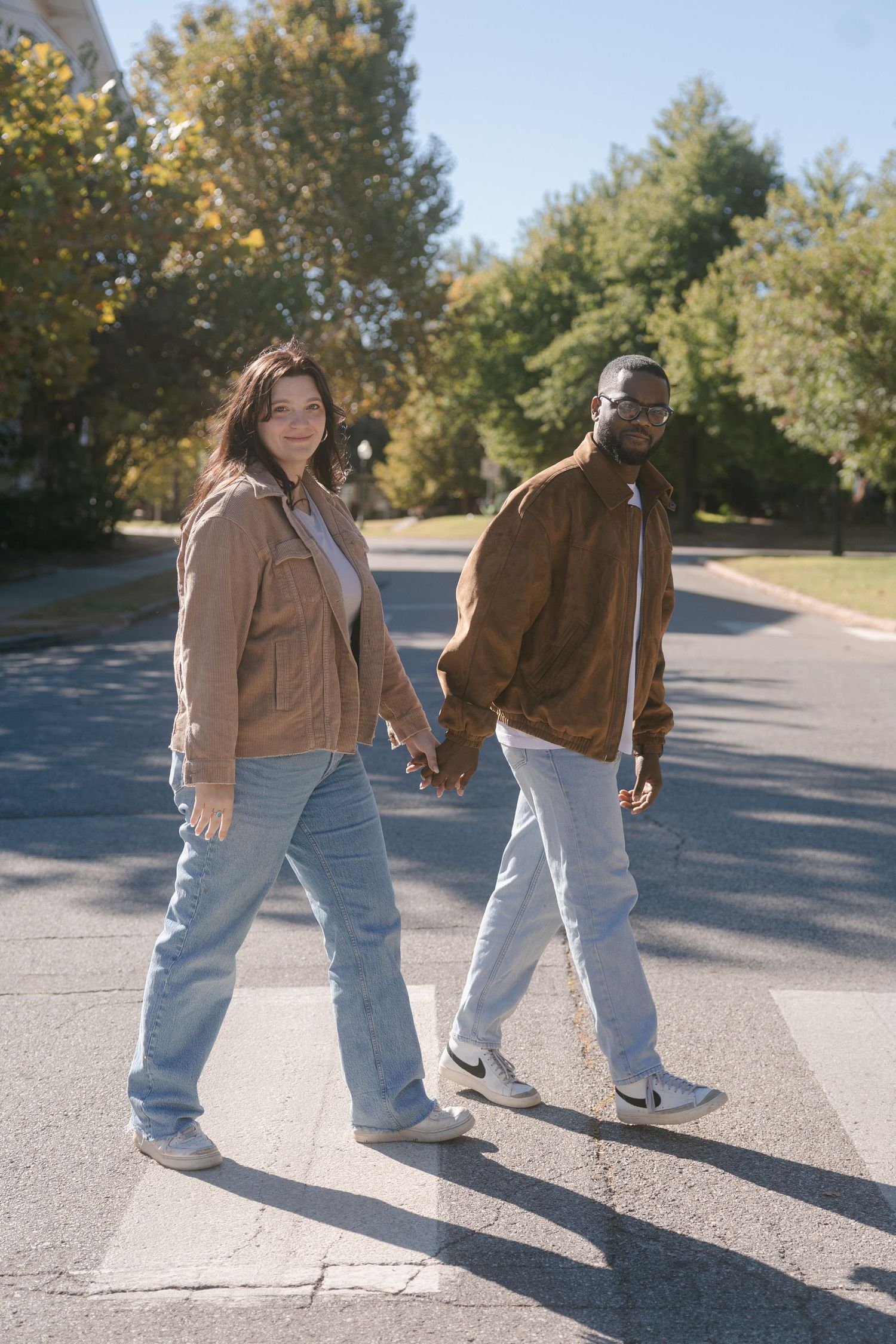 A couple wearing matching brown jackets and jeans hold hands while walking together on a sunny autumn day.