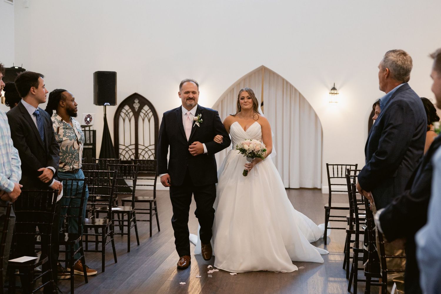 Wedding ceremony guests stand and watch as the bride walks down the aisle in an elegant white venue.