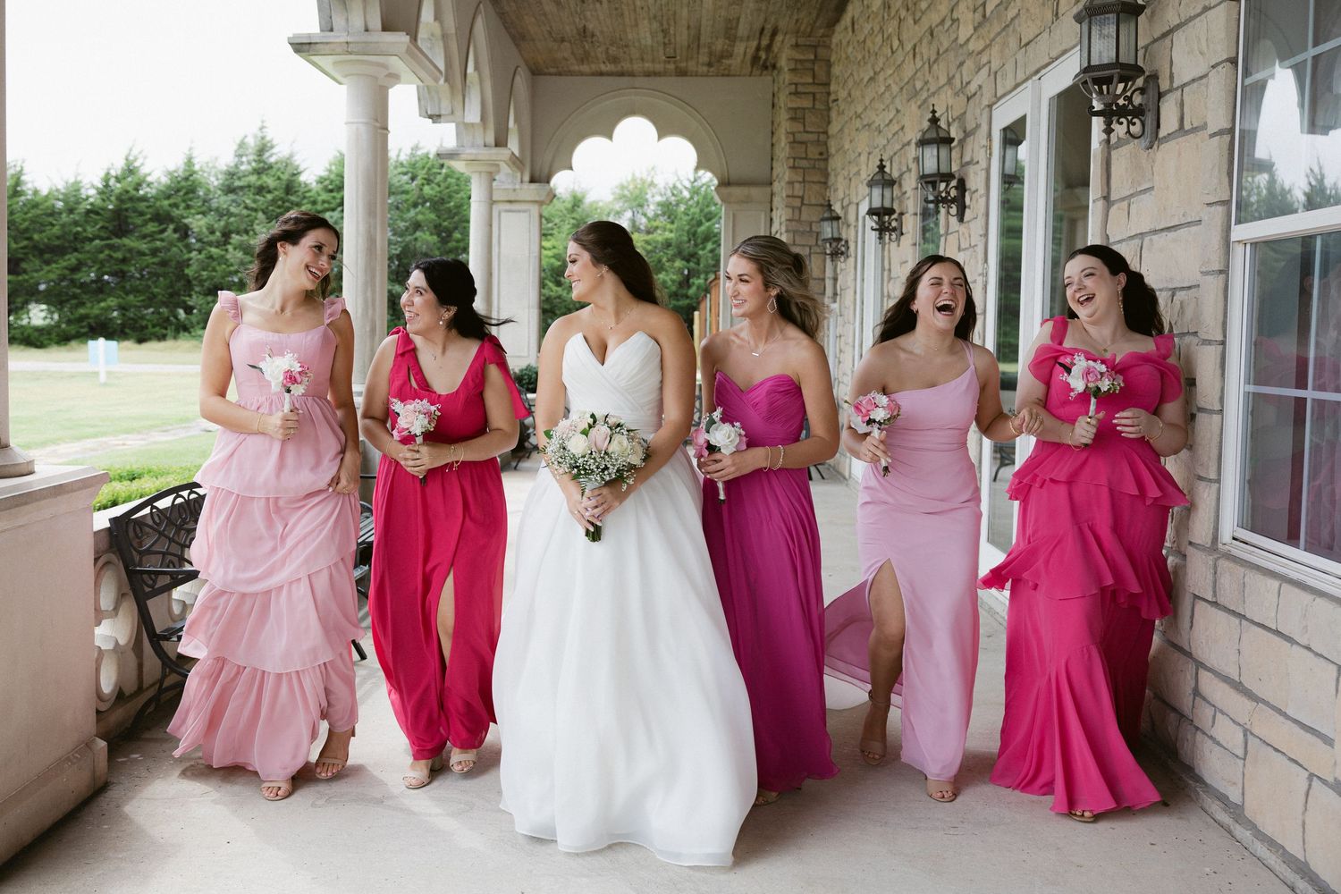 A wedding party poses in pink and fuchsia bridesmaid dresses with the bride in white on a covered porch.