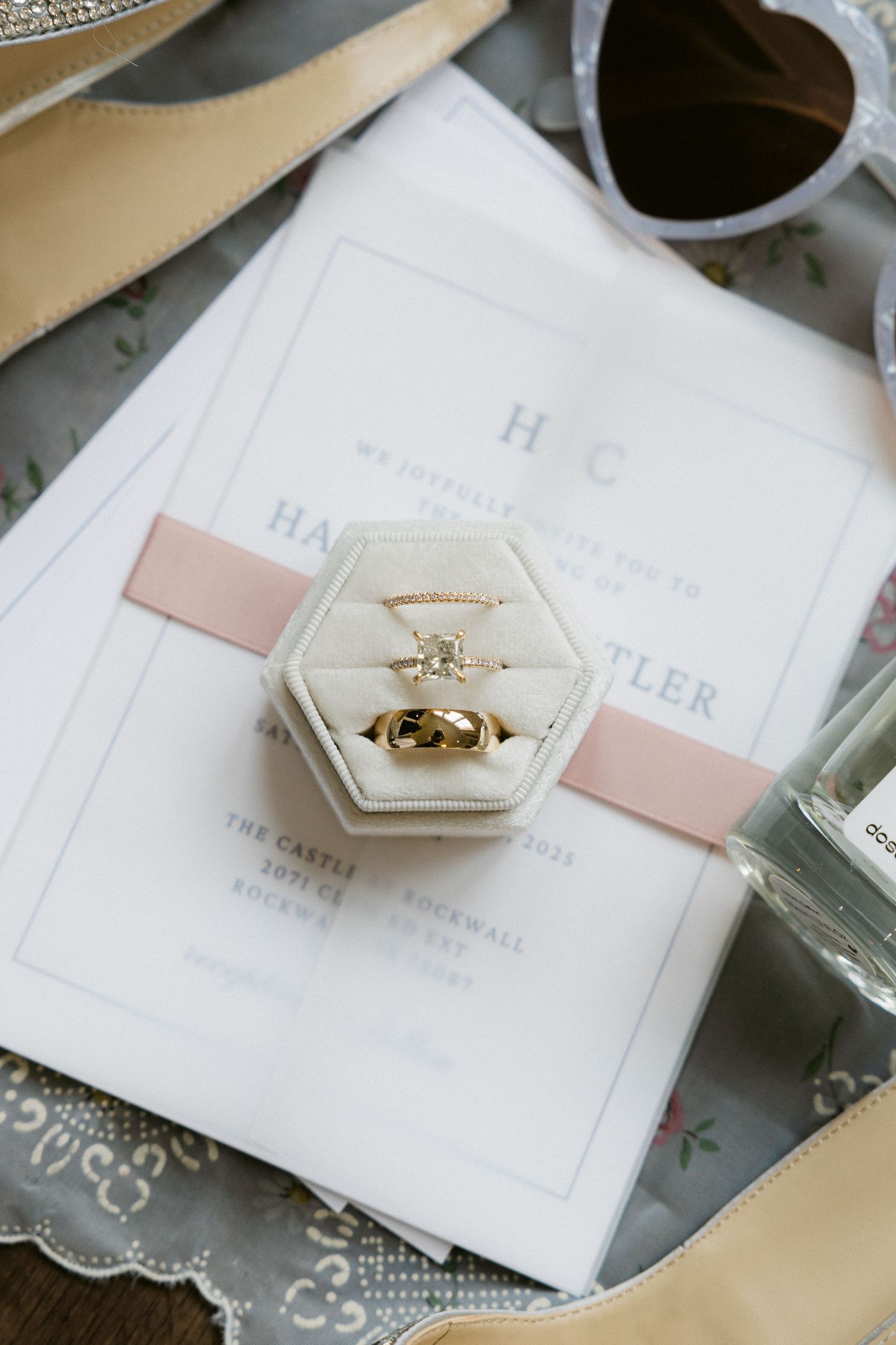 Wedding rings displayed in a hexagonal velvet ring box with pink ribbons on white paper and sunglasses nearby.