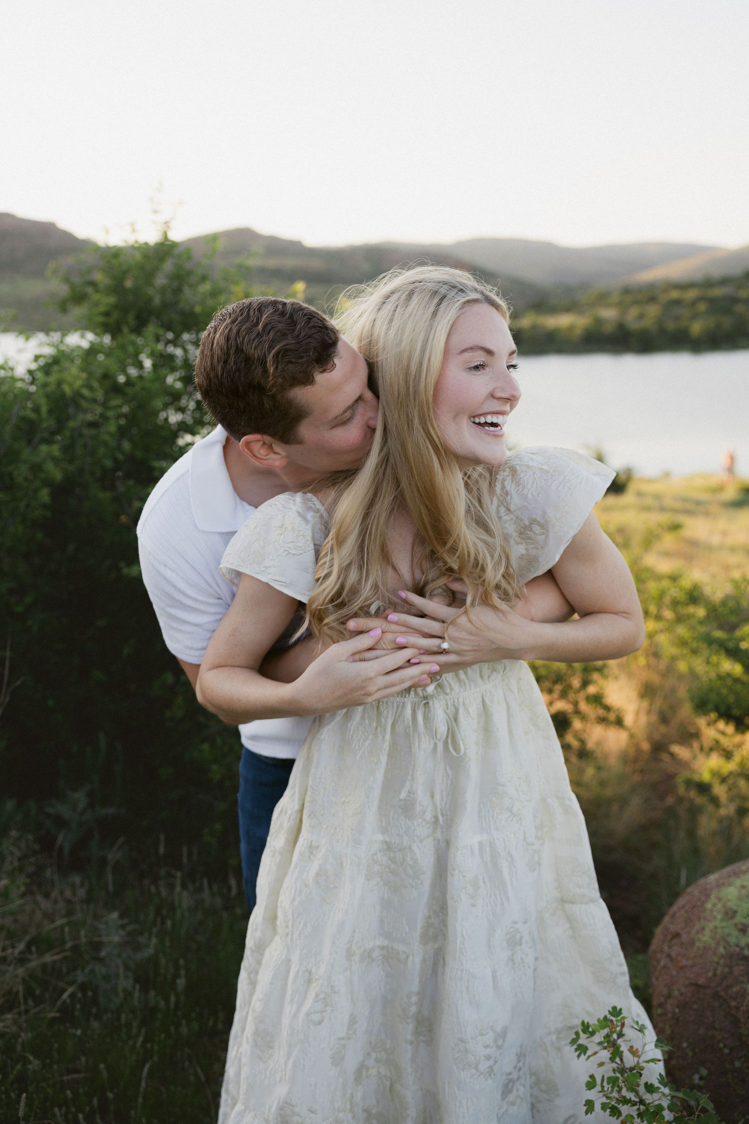 A romantic couple in white clothing embraces and laughs together in a scenic outdoor setting at golden hour.