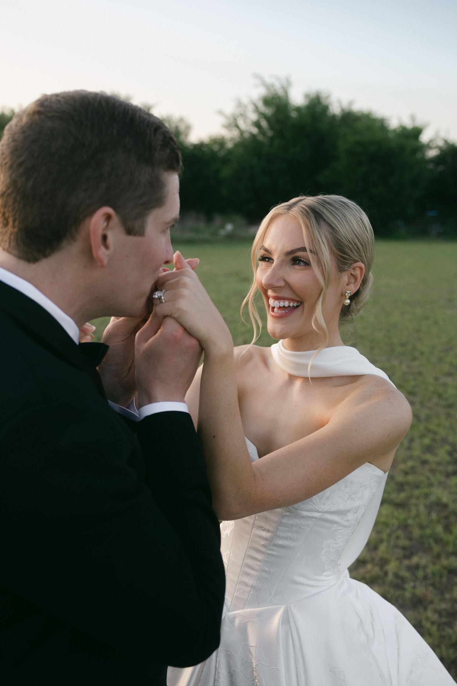 Groom kissing bride's hand as she laughs joyfully in white gown during outdoor portrait.