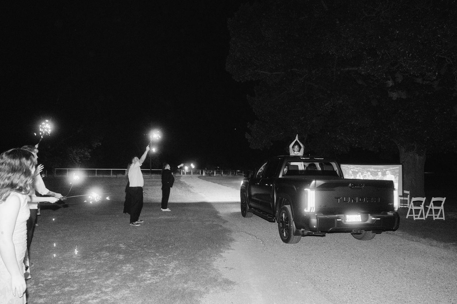 Black and white nighttime photo of people holding sparklers near a dark truck on a rural path.