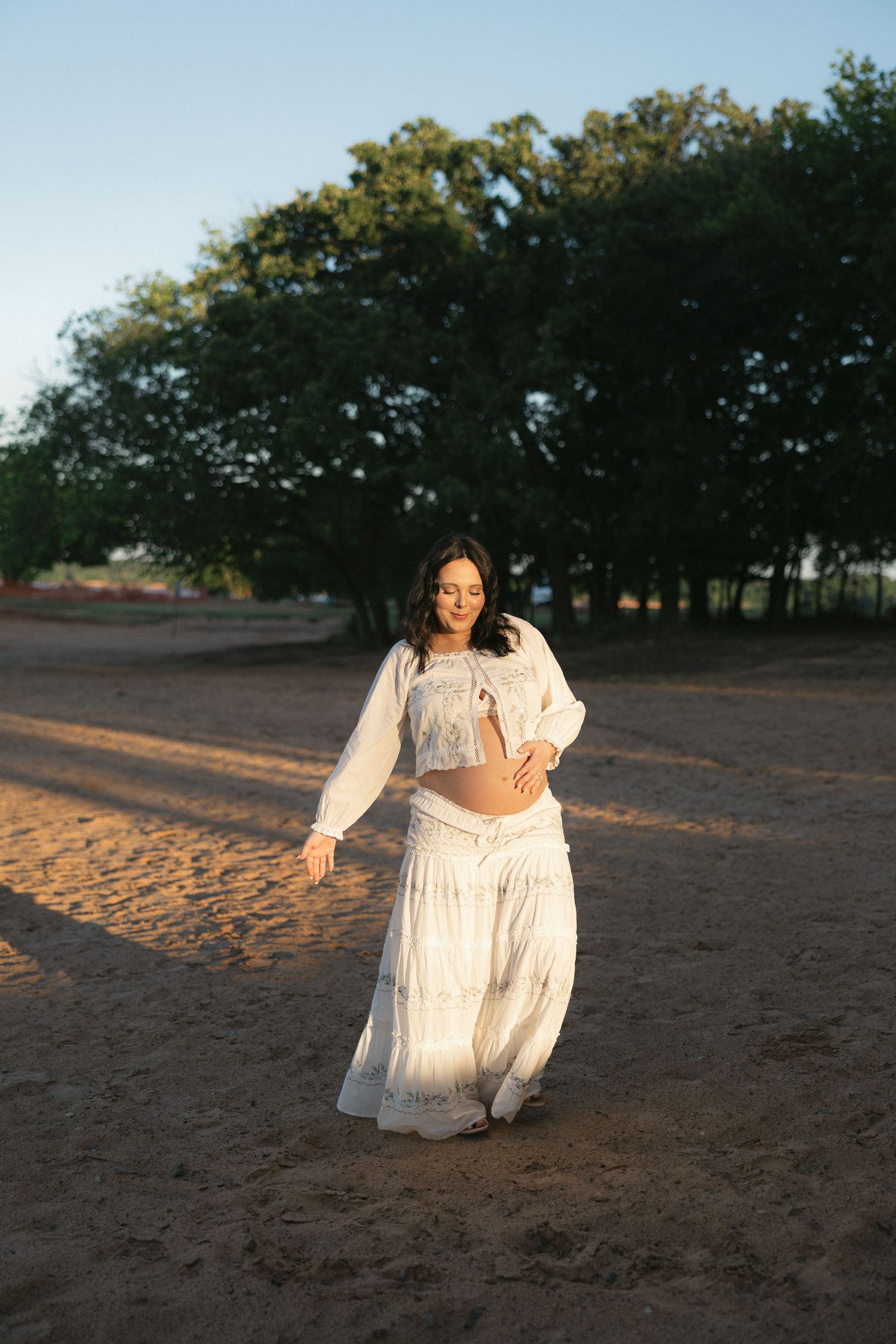 Pregnant woman in white crop top and flowing skirt standing on sandy ground with trees in golden hour light.