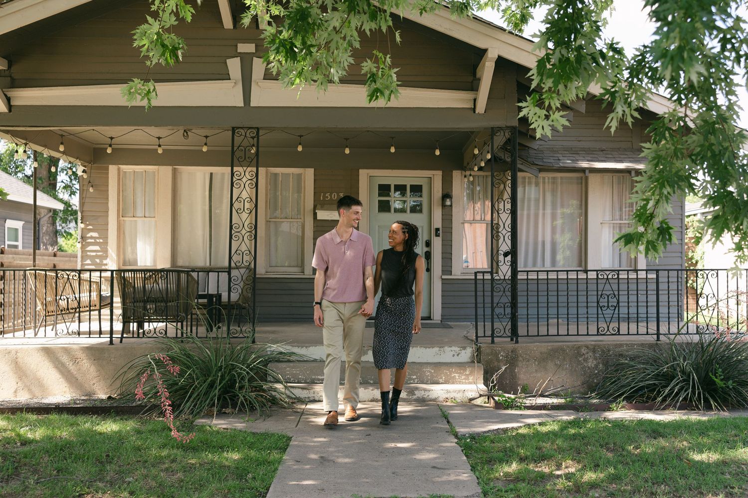 Happy interracial couple walking hand-in-hand along a tree-lined path toward a craftsman bungalow.