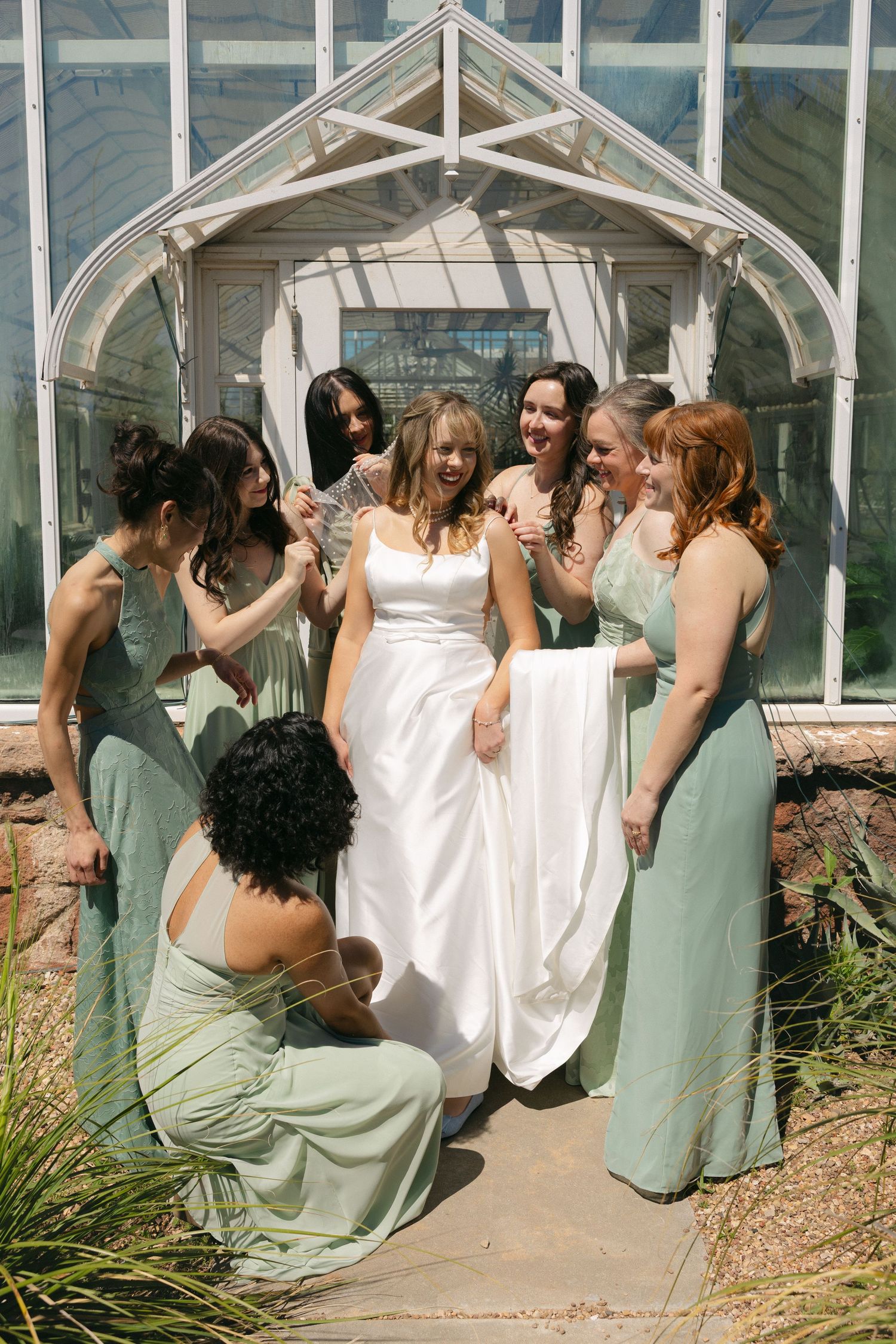 Bride in white gown surrounded by bridesmaids in sage green dresses outside a greenhouse venue.