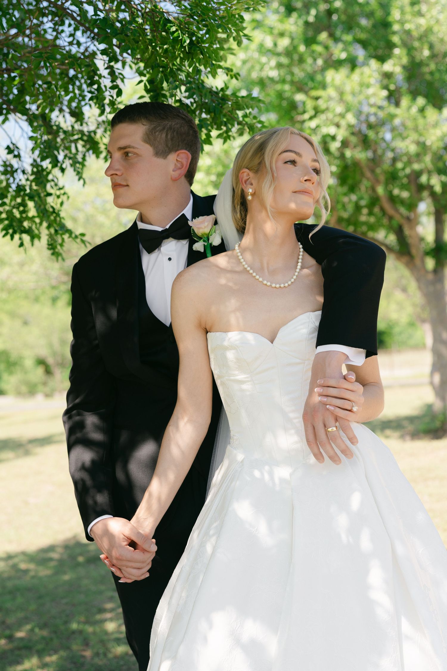Bride in white strapless gown and groom in black tuxedo hold hands outdoors under green trees on wedding day.