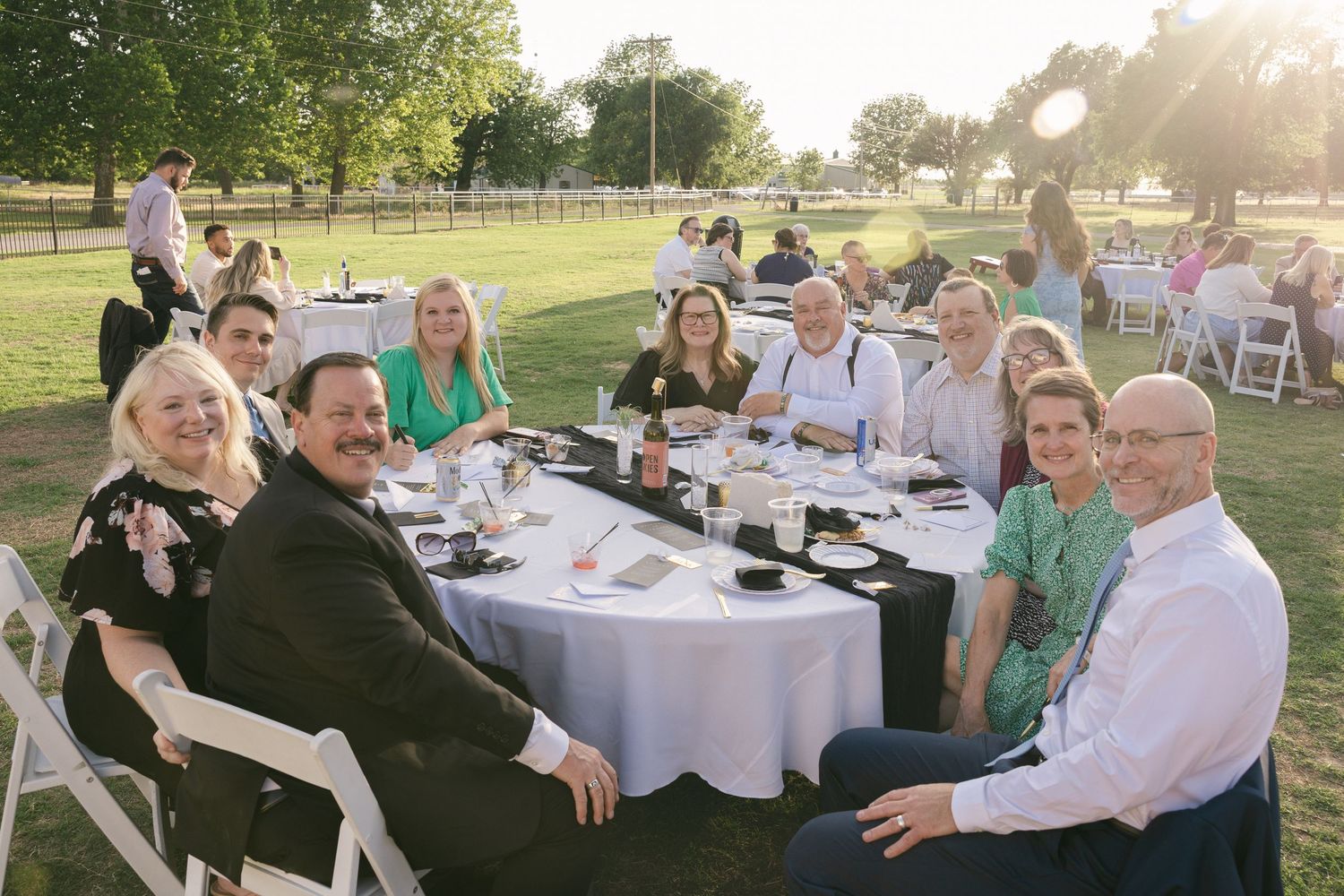 Group of elegantly dressed guests smiling at a round table during an outdoor evening wedding reception on green lawn.