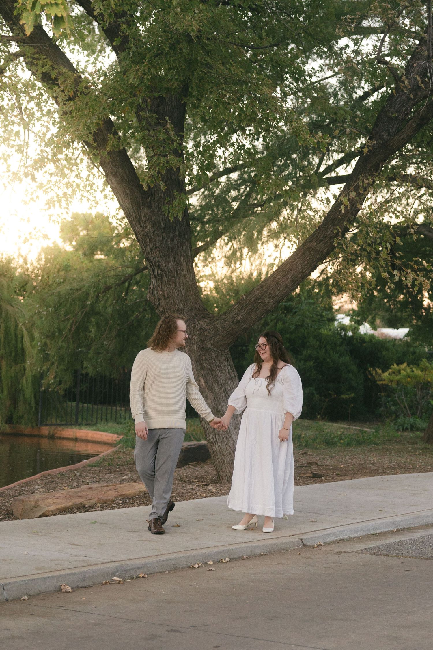 A couple sits and walks together on a concrete path surrounded by lush greenery and trees at sunset.