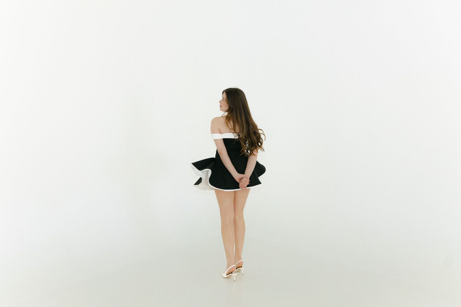 Young woman in black mini skirt and white off-shoulder top posing gracefully against white background.