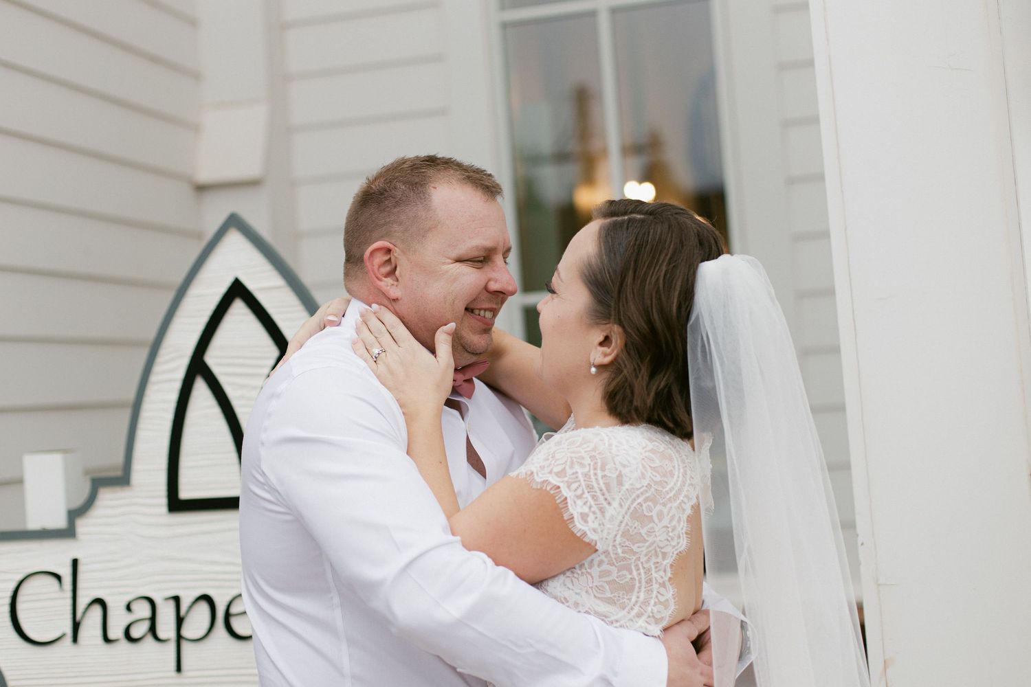A couple in wedding attire shares a romantic kiss in front of a white chapel wall.
